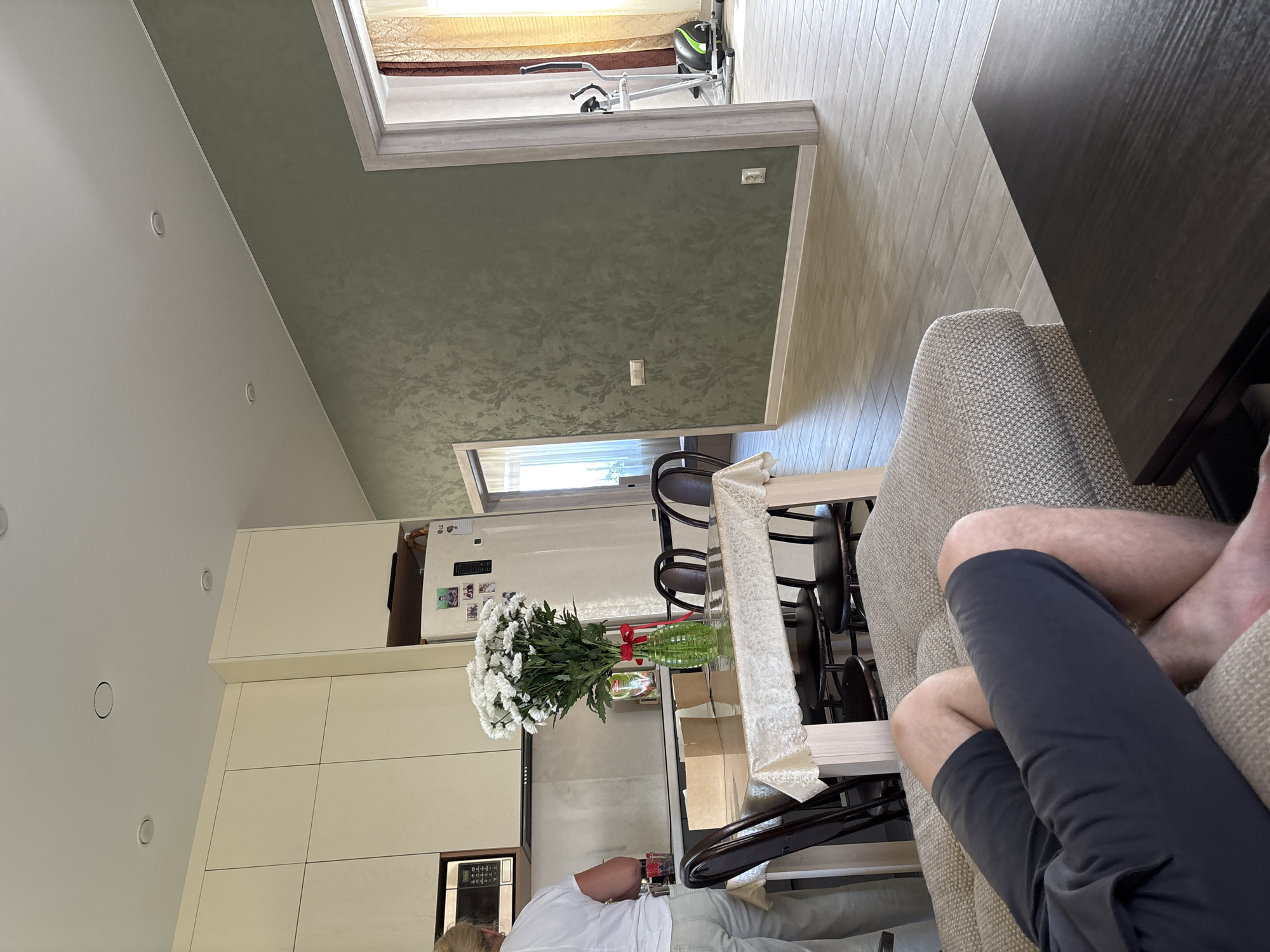 The image shows an interior view of a home, potentially in Kochkino, Russia, featuring a partial view of a person sitting on a light-colored sofa wearing dark gray shorts. Adjacent to the sofa is a dark wooden table. A dining area includes dark-colored chairs and a table covered with a white lace tablecloth. A large bouquet of white flowers sits on the table in a green vase with a red ribbon. A partial view of another person wearing a white shirt and gray pants is visible near the dining table.
The environment includes a modern kitchen with light-colored cabinets and a refrigerator. A door/window frame is seen in the background, leading to an area with a fitness bike. The walls are painted in two different colors: a lighter shade in the kitchen area and a textured, darker shade above. The flooring appears to be wooden. Lighting is provided by recessed ceiling lights.