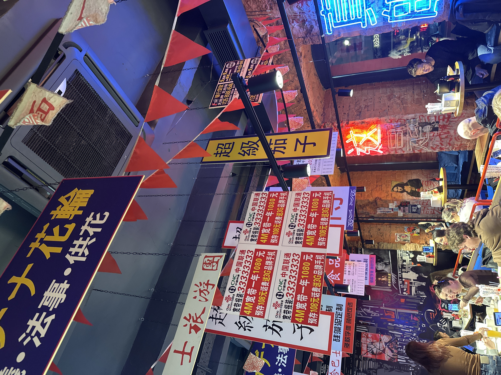 The image shows an interior space in what appears to be a restaurant or eatery. The setting is cluttered and features an array of banners and signs in Chinese. Patrons are seated at tables, with some appearing to be eating or using their phones. Neon signs adorn the walls, and a variety of hanging decorations such as pennant flags and lighting fixtures are visible. The decor suggests an attempt to evoke a lively, possibly nostalgic, atmosphere reminiscent of a street food market. The scene appears to be vibrant and populated. There is an air conditioning unit visible on the top left of the image.