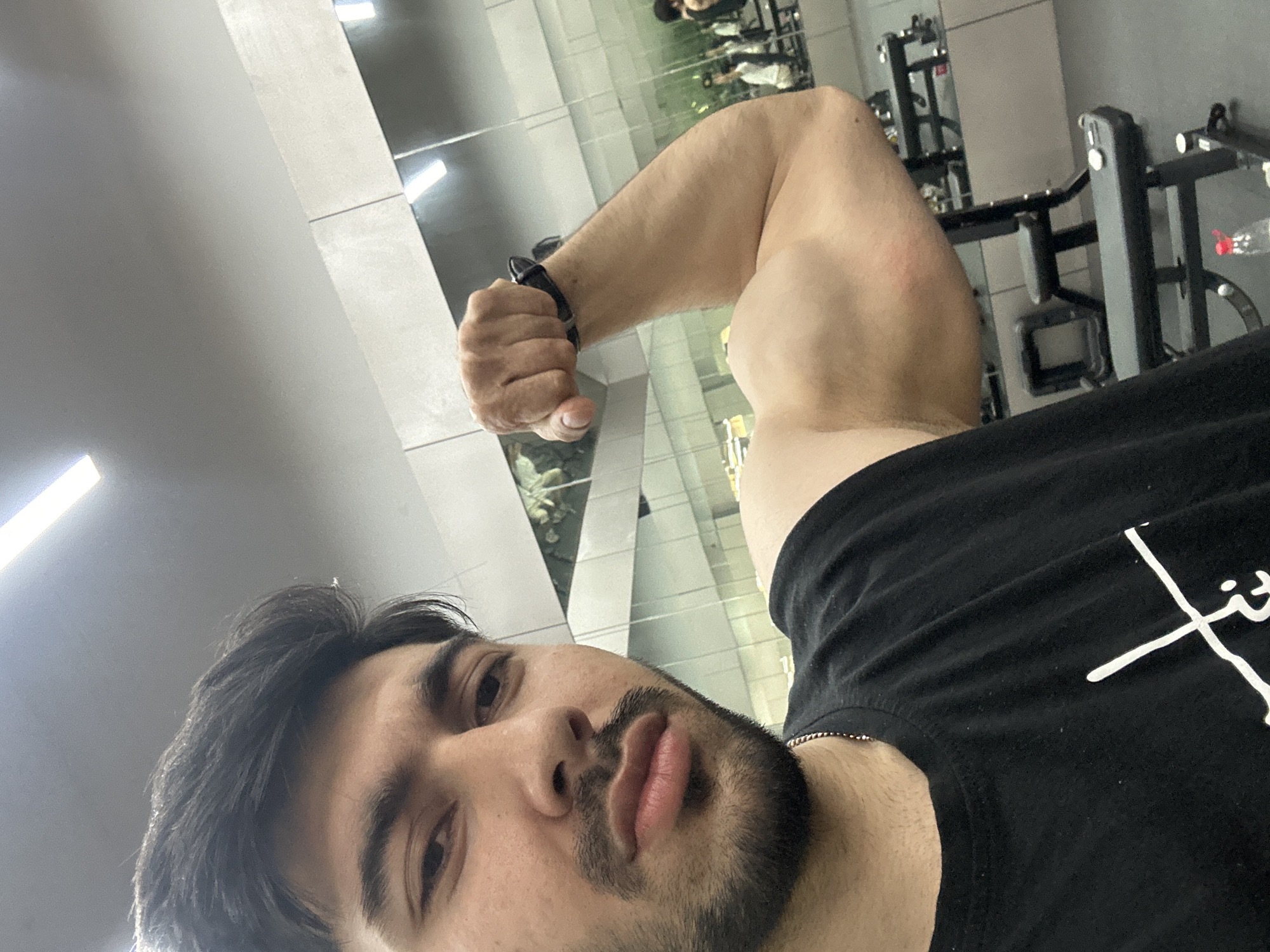 A young man takes a selfie while flexing his left bicep in what appears to be a gym or fitness center located in Chongqing High-tech Zone, China. The image is taken from a slightly upward, close-up angle, focusing on the man's face and arm.
The man has dark hair, a neatly trimmed beard and mustache, and dark eyes. He wears a black t-shirt with a partial white graphic that seems to spell out "FITT" or a similar brand name, and a thin silver chain around his neck. A dark-colored smartwatch or wristband is visible on his left wrist as he makes a fist, showcasing his bicep. His expression is neutral and focused.
The setting is an indoor space with bright overhead lighting. The background features large mirrored walls, reflecting gym equipment such as racks and machines, as well as the blurry figures of other people working out. A water bottle with a red cap is faintly visible in one of the reflections. The floor and ceiling are light-colored, possibly concrete or tiled, with linear light fixtures. The overall atmosphere suggests a modern fitness facility.