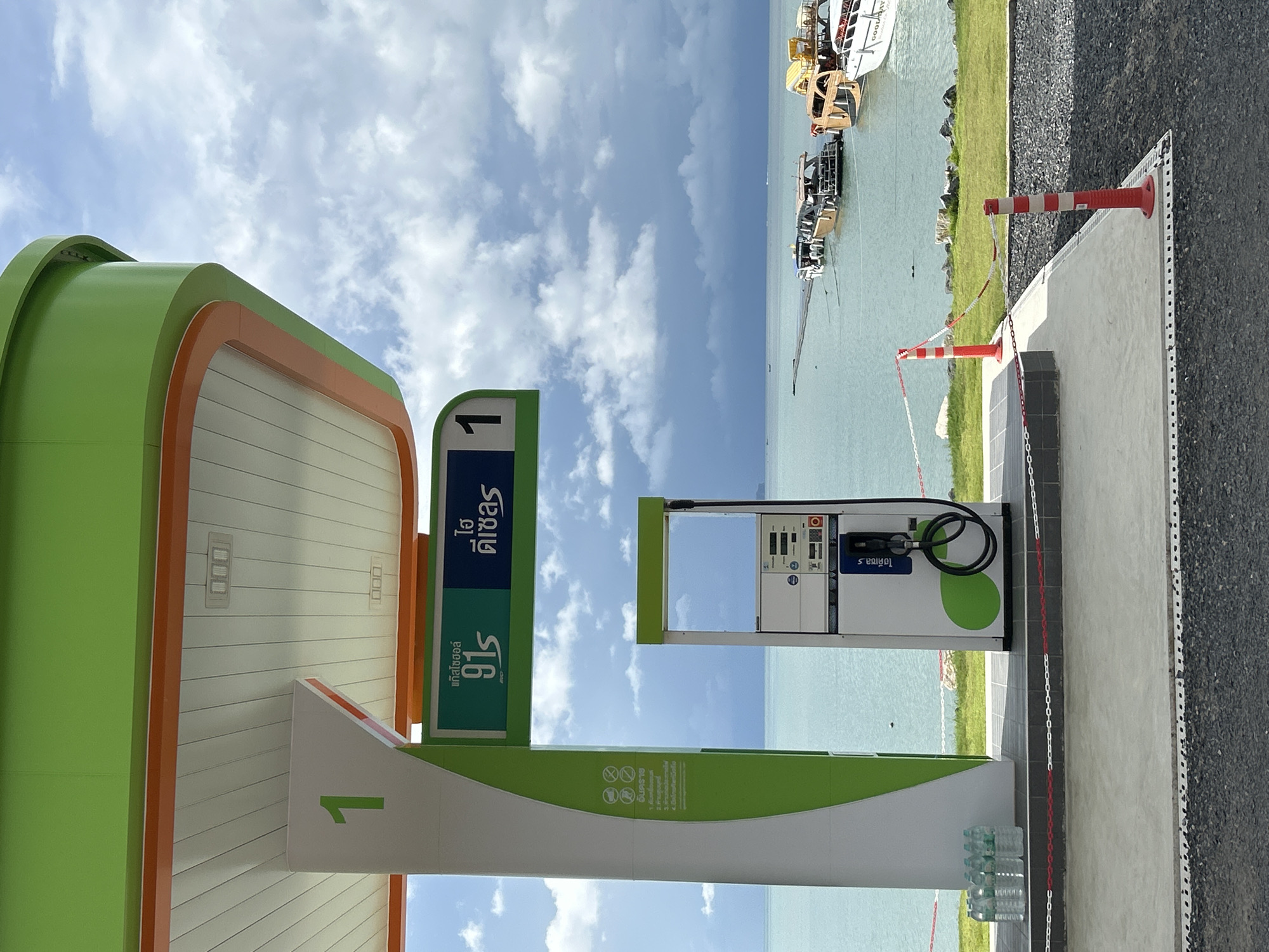 The image depicts a modern, brightly colored gas station canopy with green and white accents, situated in Ratsada, Thailand. The canopy covers a single gas pump, which is white with green circular accents and a black hose. To the left of the pump, a large white pillar displays the number "1" in green. Below this pillar, several bottles of water are neatly arranged. A green sign above the pump indicates fuel types in Thai script.
The gas station is positioned adjacent to a body of water, likely the Andaman Sea, given its location in Thailand. The water is a tranquil blue-green, and several boats, including speedboats and a larger vessel with a wooden hull, are docked or moored in the distance. A wooden walkway extends into the water. The shoreline is lined with green grass and a low wall. The sky is partially cloudy, with dramatic, dark clouds in the distance contrasting with lighter, scattered clouds overhead. Red and white stanchions with chain barriers mark the perimeter of the paved area. The overall impression is one of a clean, well-maintained, and picturesque service station by the sea.