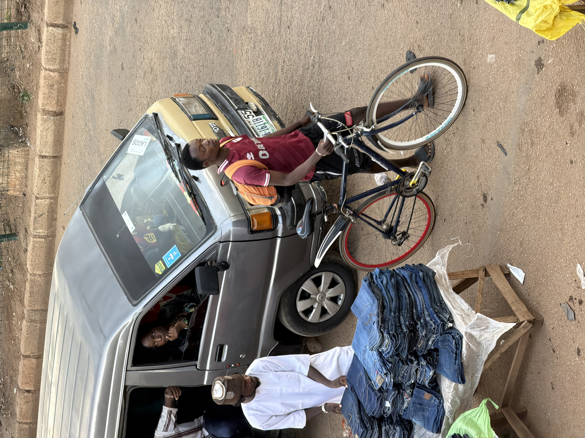 A street scene in Bukuru, Nigeria, features a silver and gold Toyota van parked on a dirt road. A Black man wearing a white robe and cap stands next to a table laden with stacks of blue jeans, appearing to be a vendor. Beside him, a young Black man with an orange backpack and a maroon and white jersey walks with a blue bicycle. A Black woman is visible inside the van, looking out the window. The scene suggests a bustling market or roadside commerce.