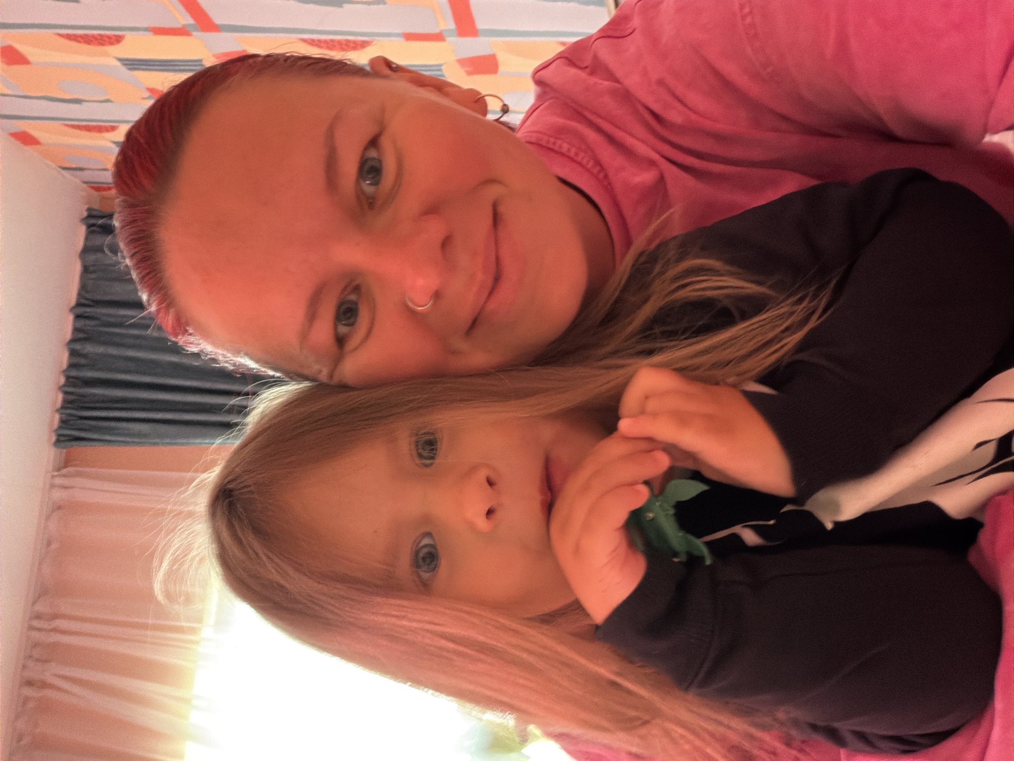 A selfie-style photograph shows two individuals. The upper individual appears to be an adult with red hair, wearing a pink top and a nose ring. The lower individual appears to be a child with blonde hair, wearing a black top. The child's hands are visible. The background shows a wall with a striped pattern and a curtain. There is not enough information to confirm the location of the photo in the city of Chaikovsky, Russia.