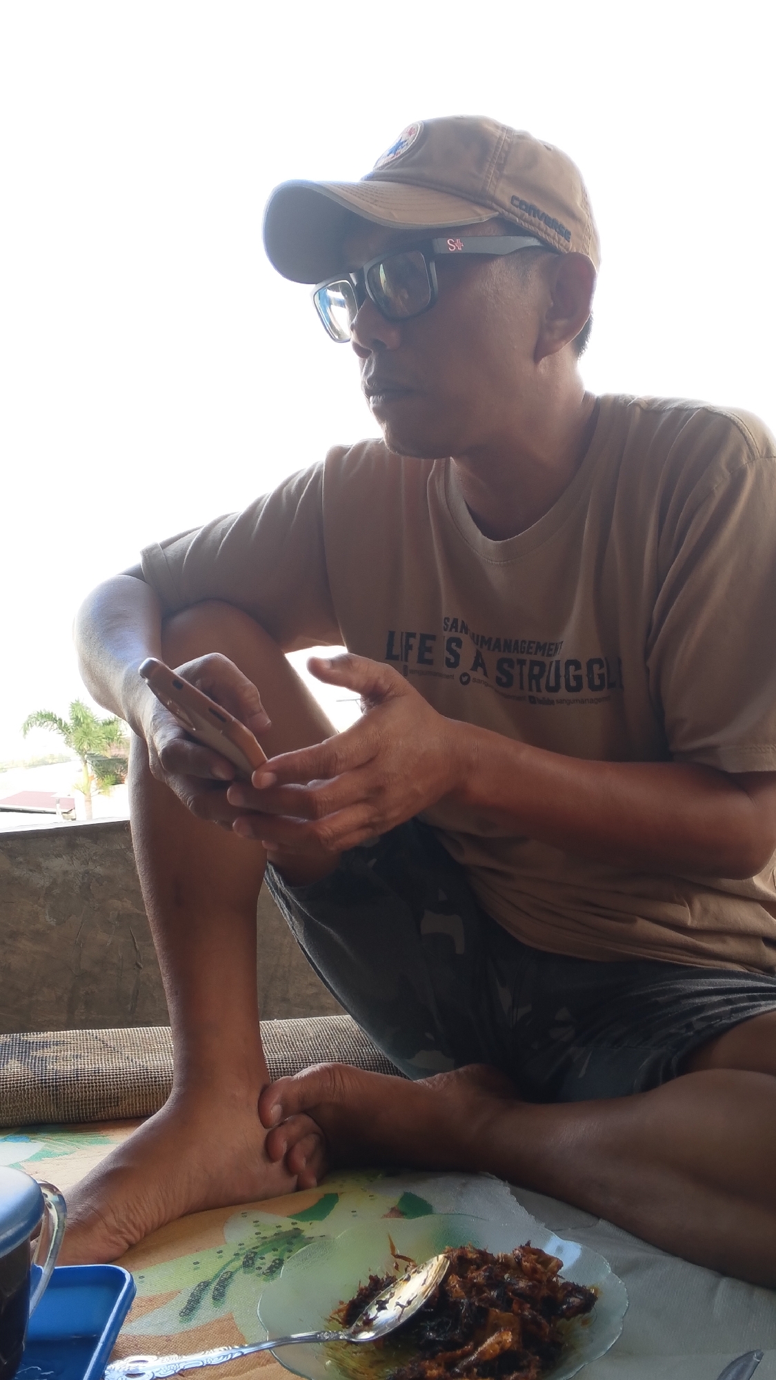 A man in a brown t-shirt and a beige baseball cap sits cross-legged on the floor, looking to his right. He wears black-rimmed glasses and is holding a smartphone in his left hand. In front of him is a plate of food with a spoon. The background is bright white, suggesting an outdoor setting with bright light. The lower portion of the image shows a patterned floor and a glimpse of outdoor scenery with a palm tree. This scene appears to be in Kabonena, Indonesia, given the contextual information.