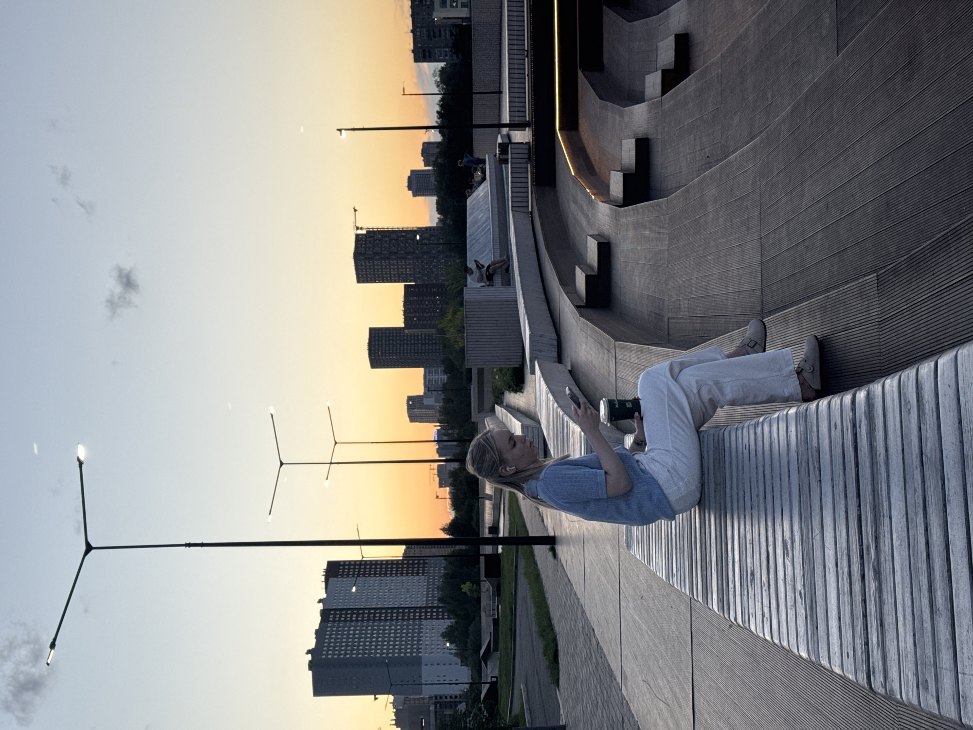 A young blonde woman with white AirPods in her ears sits on a wooden bench, looking at her smartphone. She is wearing a light blue short-sleeved shirt and white pants. In her right hand, she holds a disposable coffee cup with a dark lid. The bench is part of a tiered outdoor seating area with wooden planks and concrete elements, illuminated by subtle lighting. In the background, a city skyline is visible with modern high-rise buildings against a gradient sunset sky, transitioning from orange to pale blue. Streetlights with multiple lamps stand tall along a paved walkway. This scene is located in the city of Yekaterinburg, Russia.