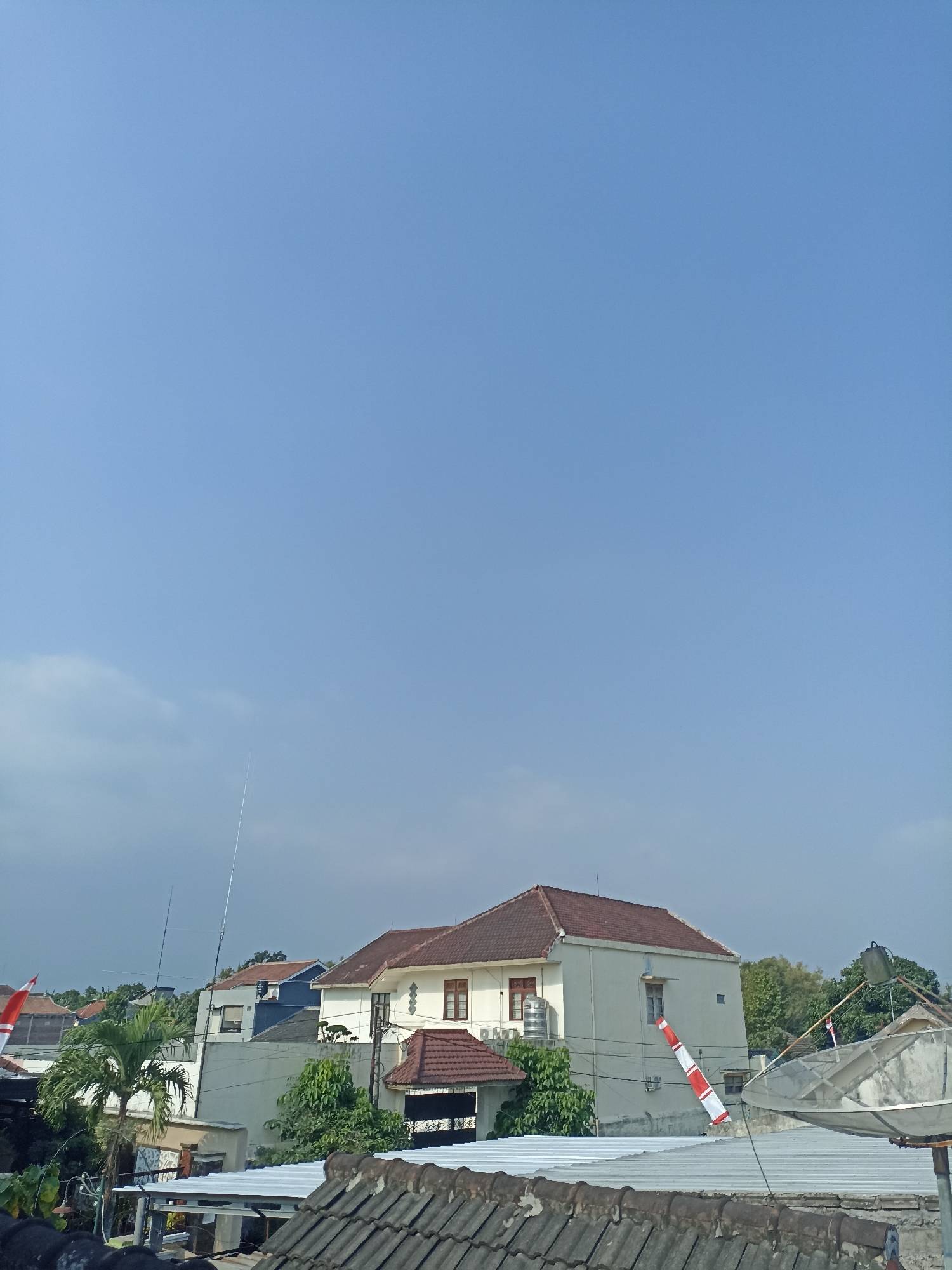 The image depicts a clear blue sky above a suburban residential area in Sumberejo, Indonesia. In the foreground, a gray tiled roof is visible, with a white corrugated metal roof and a satellite dish to its right. Further back, a light-colored, multi-story house with a brown tiled roof stands out. To its left, smaller houses and lush green trees are present. Two tall, slender antenna masts are visible, one taller than the other. A red and white striped windsock hangs from the side of the main house. On the far left, a flag with red and white stripes, likely the Indonesian flag, is partially visible. The scene suggests a sunny day in a residential neighborhood.