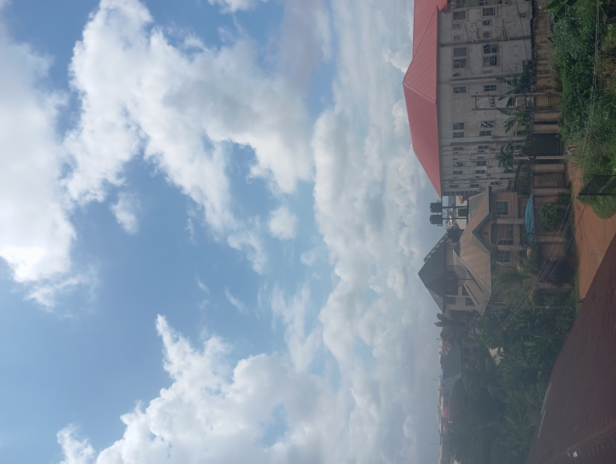 This is an eye-level, full exterior shot of a residential area in Nkpor, Nigeria, under a bright, cloudy sky. In the foreground, a dark, corrugated metal roof slopes from left to right. Beyond this, a dirt road curves into the middle ground, flanked by lush green vegetation and trees. Several buildings are visible, ranging from multi-story houses with metal roofs to larger structures with red and grey tiled roofs. One prominent building on the right is a large, light-colored structure with a red roof, featuring numerous windows and an attached water tank system. In the background, a dense cluster of residential buildings and trees stretches across the horizon. Power lines and poles are interspersed throughout the scene. No people or animals are discernible in the image. The overall impression is a view of a densely populated urban or suburban environment.