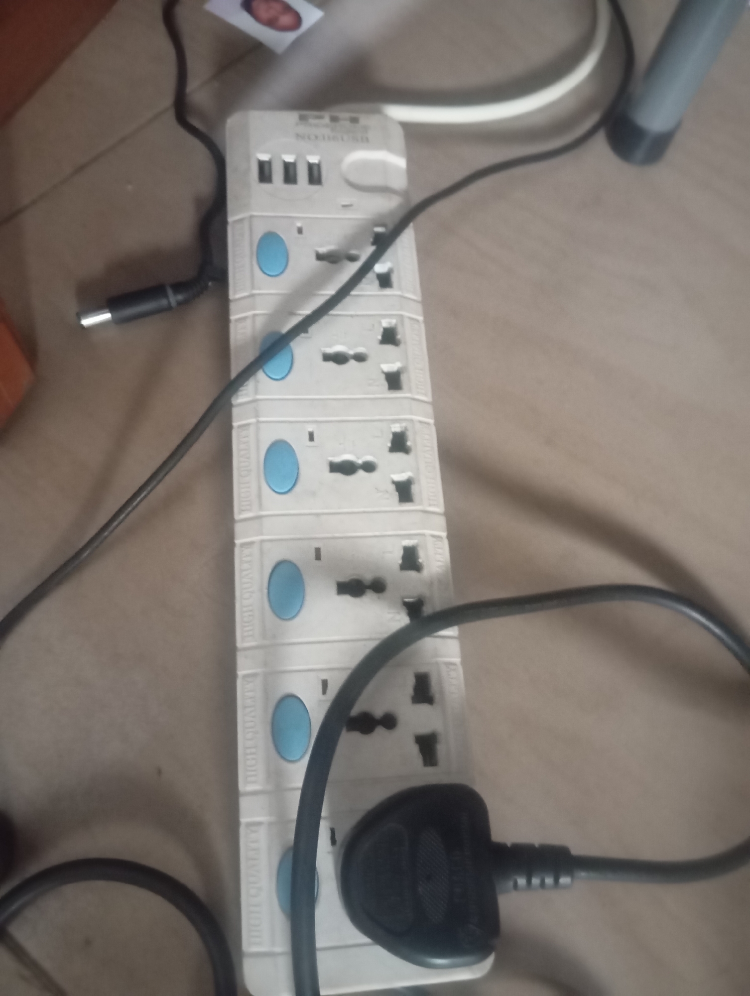 The image shows a power strip with multiple outlets, connected to a power cord with a plug. The power strip is white and has several blue circular buttons, possibly indicating individual switches for each outlet. There are also three USB ports at the top of the power strip. The power strip is lying on a tiled floor. A black cable from a device is plugged into one of the outlets. The image also shows a small picture of a person's face in the upper left corner. The scene appears to be indoors. The request for the location in Jos, Nigeria cannot be fulfilled as there is no visual information in the image to support this specific detail.