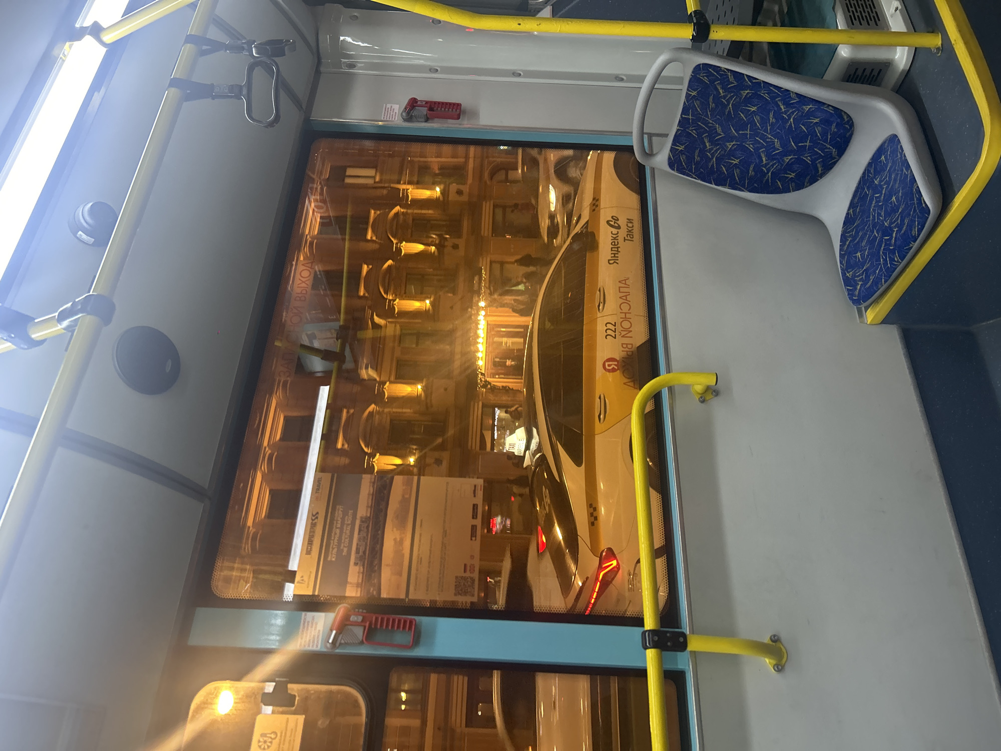 The image captures an interior view of a public transport vehicle, likely a bus or tram, at night in Saint Petersburg, Russia. The perspective looks outwards through a large window, simultaneously reflecting parts of the interior and revealing the urban street scene outside.
Inside the vehicle, bright yellow grab rails are prominent, with a dark grey handle for standing passengers. A light-colored seat with a distinctive blue and yellow abstract pattern is visible on the right. A red emergency brake or hammer is mounted below the window. Overhead lights and a speaker are also visible on the ceiling.
Through the window, a bright yellow and white car, identified as a "Yandex Go Такси" (Yandex Go Taxi) with the number "222" and the phrase "Я В ОНЛАЙНЕ" (I'm online) in Russian, is seen alongside. The exterior scene is brightly lit by streetlights and interior lights of buildings, showcasing ornate facades typical of Saint Petersburg.
The window's reflective surface also displays text from interior signs or stickers, including "КУЛЬТУРНЫЙ МАРШРУТ" (Cultural Route), "THE CULTURAL ROUTE", and "TRAVEL", along with QR codes. The word "ВЫХОД" (Exit) is visible in reverse, indicating a sign inside the bus. The scene depicts a moment of urban transit, with artificial lights illuminating both the interior and the nighttime city environment. No individuals are explicitly visible.