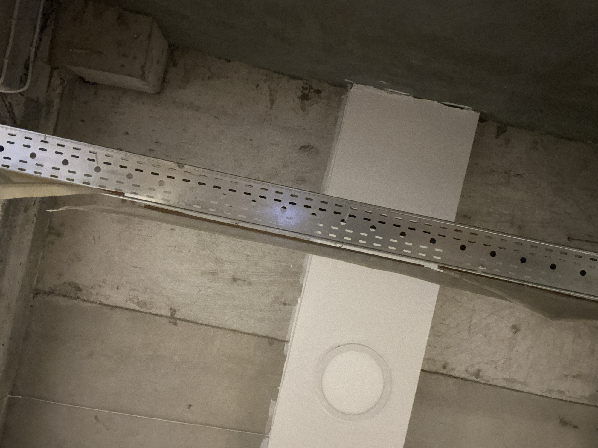 A low-angle, indoor shot depicts a concrete ceiling with a rectangular white object that appears to be a light fixture. A metal cable tray, perforated with holes, runs diagonally across the center of the frame. The surrounding concrete ceiling shows signs of age or wear, with some discoloration and rough texture. The overall setting appears to be in an unfinished or industrial space.