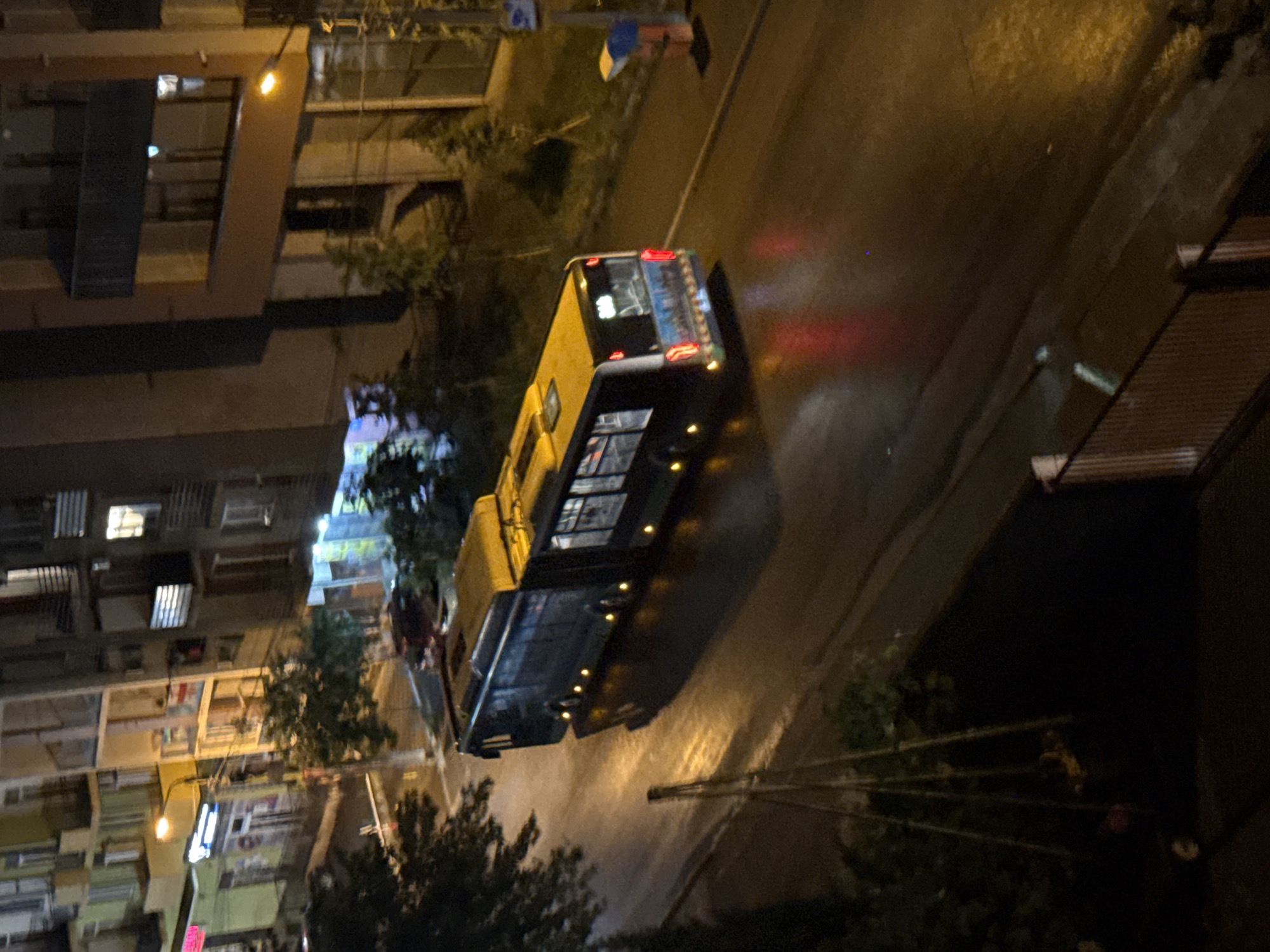 An articulated bus, dark-colored with a yellow roof, travels on a wet asphalt road at night. The bus's interior lights are illuminated, and its exterior lights, including headlights and taillights, are visible. The bus is in motion, casting reflections on the wet road. In the background, multi-story buildings line the street, with some windows lit. Streetlights cast a warm glow, and trees are present, partially obscuring the buildings. A metal railing is visible in the foreground, suggesting an elevated viewpoint, likely a balcony or window. The scene appears to be in an urban environment, possibly a residential or mixed-use area. The location is identified as Tbilisi, Georgia.