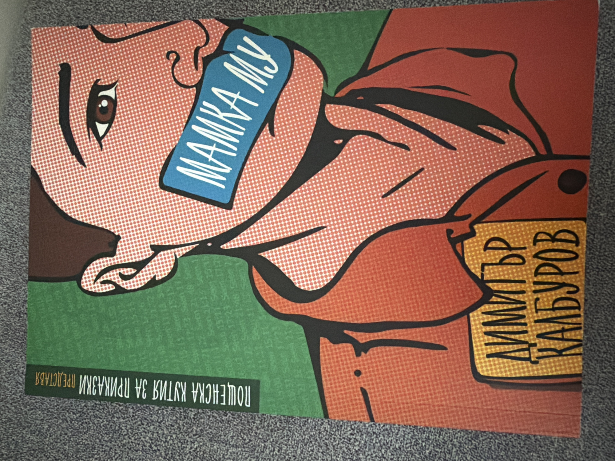 This is a photograph of a poster featuring a stylized portrait of a person with a blue band across their mouth that reads "МАМКА МУ" in white letters. The person's face is rendered in a comic-book style with orange and white dots. A green vertical band runs along the left edge of the poster with text in Bulgarian that reads "ПОЩЕНСКА КУТИЯ ЗА ПРИКАЗКИ ПРЕДСТАВЯ". Below the portrait, a yellow rectangular tag contains the name "ДИМИТЪР КАЛБУРОВ" in black lettering. The poster is displayed against a textured gray background. The poster's design and content suggest it is related to a theatrical performance or literary work. It is not possible to determine the specific location in Pazardzhik, Bulgaria from the image alone.