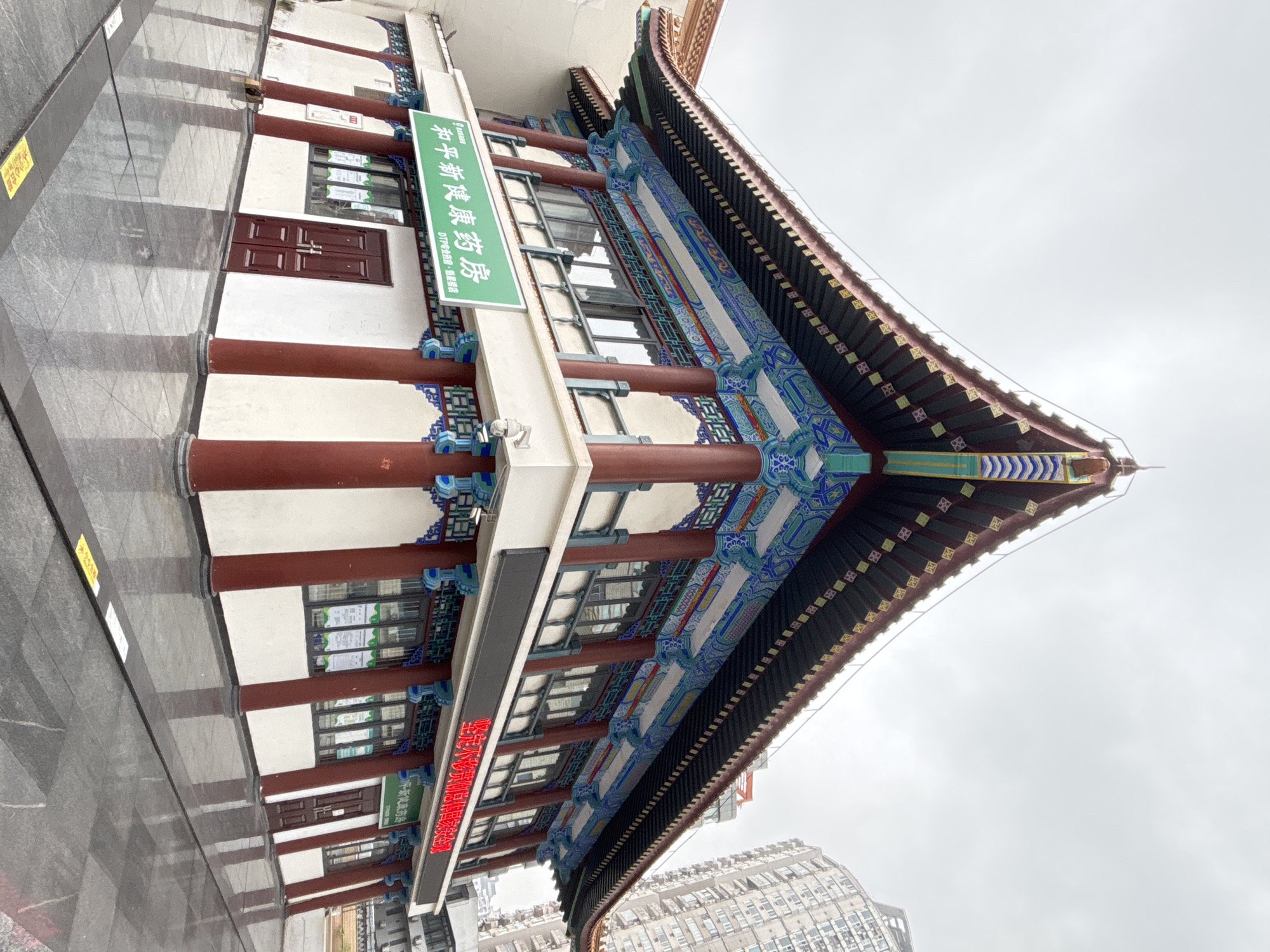 The image shows a traditional Chinese architectural structure, likely a building facade, in Yuzhong District, China. The structure has white walls, red support columns, and a multi-tiered roof with intricate blue, green, and gold detailing. Green signage with Chinese characters is visible, along with a red LED sign. The environment includes a reflective pavement area and modern high-rise buildings in the background. The sky is overcast. The focus is on the architecture and its combination of traditional and modern elements.