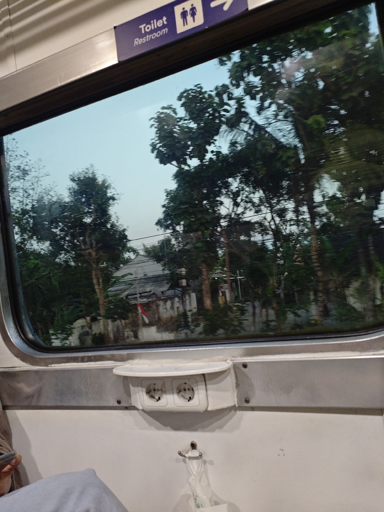 The image shows the interior of a train carriage with a view out of the window. The window displays a blurry scene of trees and buildings, with a bright, overcast sky. A sign above the window reads "Toilet Restroom" with a pictogram of two people. Inside the carriage, there are two electrical outlets mounted on the wall below the window, and a small white shelf is attached above them. A person's arm and hand, holding a smartphone, are visible in the lower left corner of the image. A plastic bag hangs from a hook on the wall. The overall scene suggests a journey by train, likely in Indonesia, given the mention of Ngadiluwih.