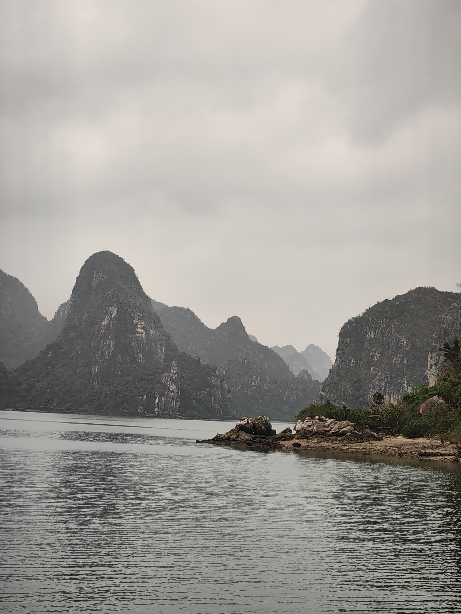 The image depicts a seascape with karst formations characteristic of Ha Long Bay, located in Bai Chay Ward, Vietnam. The foreground features calm water with horizontal ripples. A small rocky outcrop with some vegetation extends into the water from the right side of the frame. In the background, several large, forested karst islands rise from the sea. The sky is overcast and gray, creating a muted, low-contrast scene. There are no people visible.
