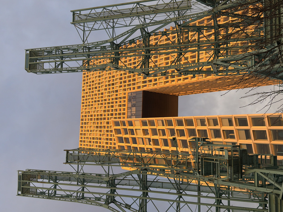 A forensic image depicts the American Copper Buildings located at 626 First Avenue in Murray Hill, Manhattan, New York City. The modern skyscraper, distinguished by its gold-toned, grid-patterned facade and a prominent rectangular archway cutting through its mid-section, is bathed in direct, warm golden light, suggesting either sunrise or sunset. The facade features numerous uniformly sized square window openings. The building is framed on both the left and right by tall, dark green metal truss structures, consistent with industrial infrastructure such as the nearby former Con Edison Kips Bay Generating Station. These structures partially obscure the building's side profiles. The sky above is a pale grey-blue with diffuse cloud cover. Bare tree branches are visible in the lower right foreground. No individuals, vehicles, or explicit actions are present in the scene.