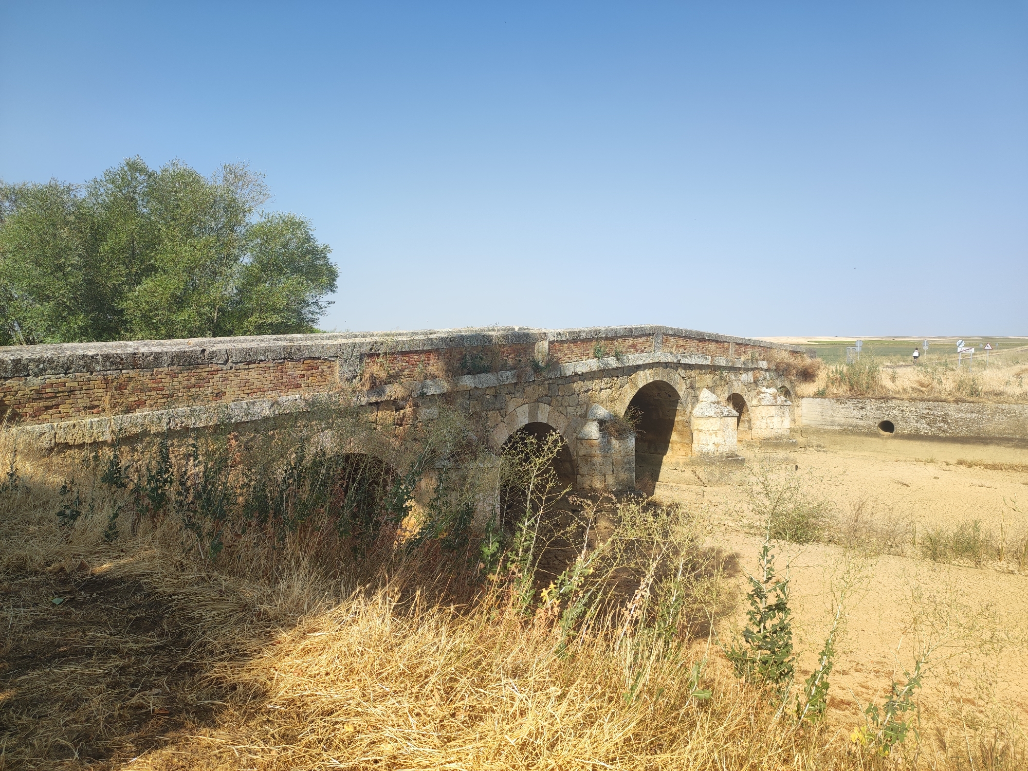 A stone and brick bridge with three visible arches spans a dry, sandy riverbed. The bridge's stone facade is weathered, with moss and vegetation growing on its lower sections. The surrounding landscape is arid, with dry grass and sparse vegetation in the foreground and a flat, open field in the background. A clear blue sky dominates the upper portion of the image. In the distance, two figures are walking on the field near a road, and several traffic signs are visible. The scene appears to be located in the rural area of Autillo de Campos, Spain, given the dry climate and traditional architecture.