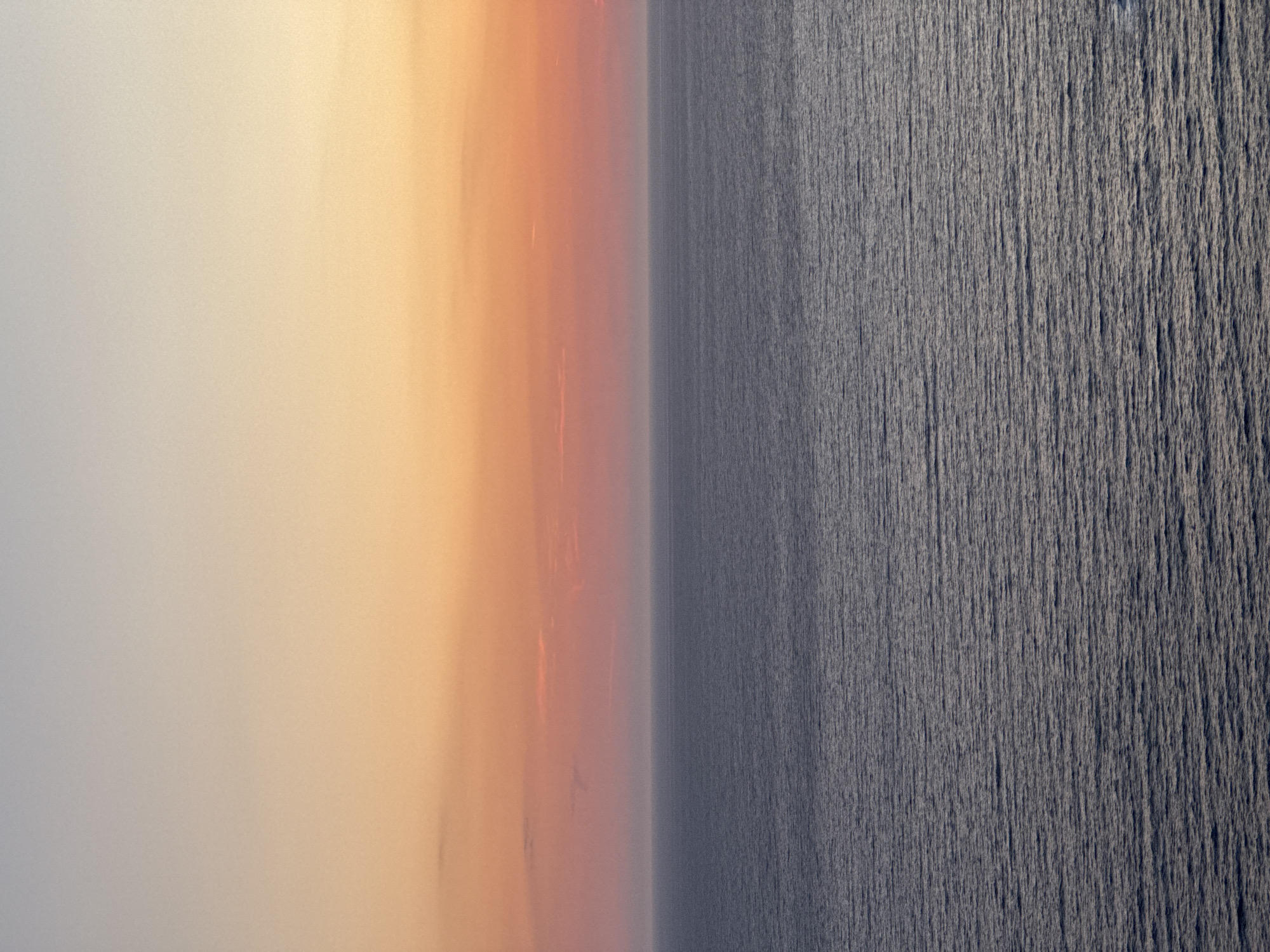 The image depicts a serene seascape at sunset, viewed from Glavatičići, Montenegro. The sky transitions from a pale, warm yellow at the top to a soft orange and pink closer to the horizon. The sea, a deep, muted blue-grey, exhibits a gentle ripple pattern, reflecting the subdued light of the setting sun. There are no people, objects, or structures visible in the frame, emphasizing the vastness and tranquility of the natural environment. The horizon line is sharp and clear, separating the sky from the ocean.