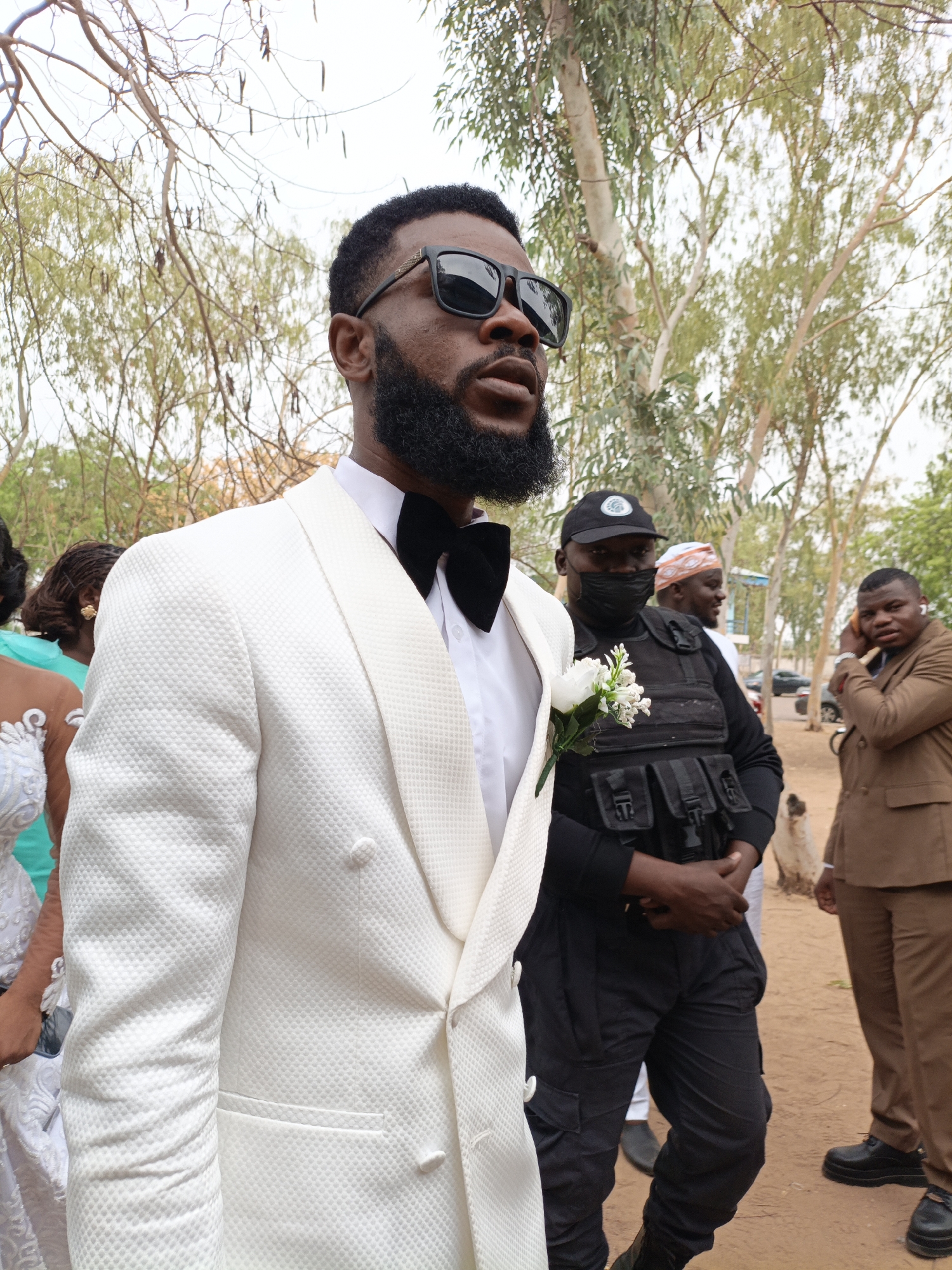 The image shows a man in a textured white suit and black bow tie standing outdoors. He wears black sunglasses and has a full beard. A white flower boutonniere is pinned to his suit. Behind him is a person in a black uniform and a mask, possibly security personnel. Another man in a brown suit is partially visible to the right. The background includes trees and sandy ground, suggesting an outdoor setting, and the location is Gawuna, Nigeria. There appears to be a wedding party nearby as well.