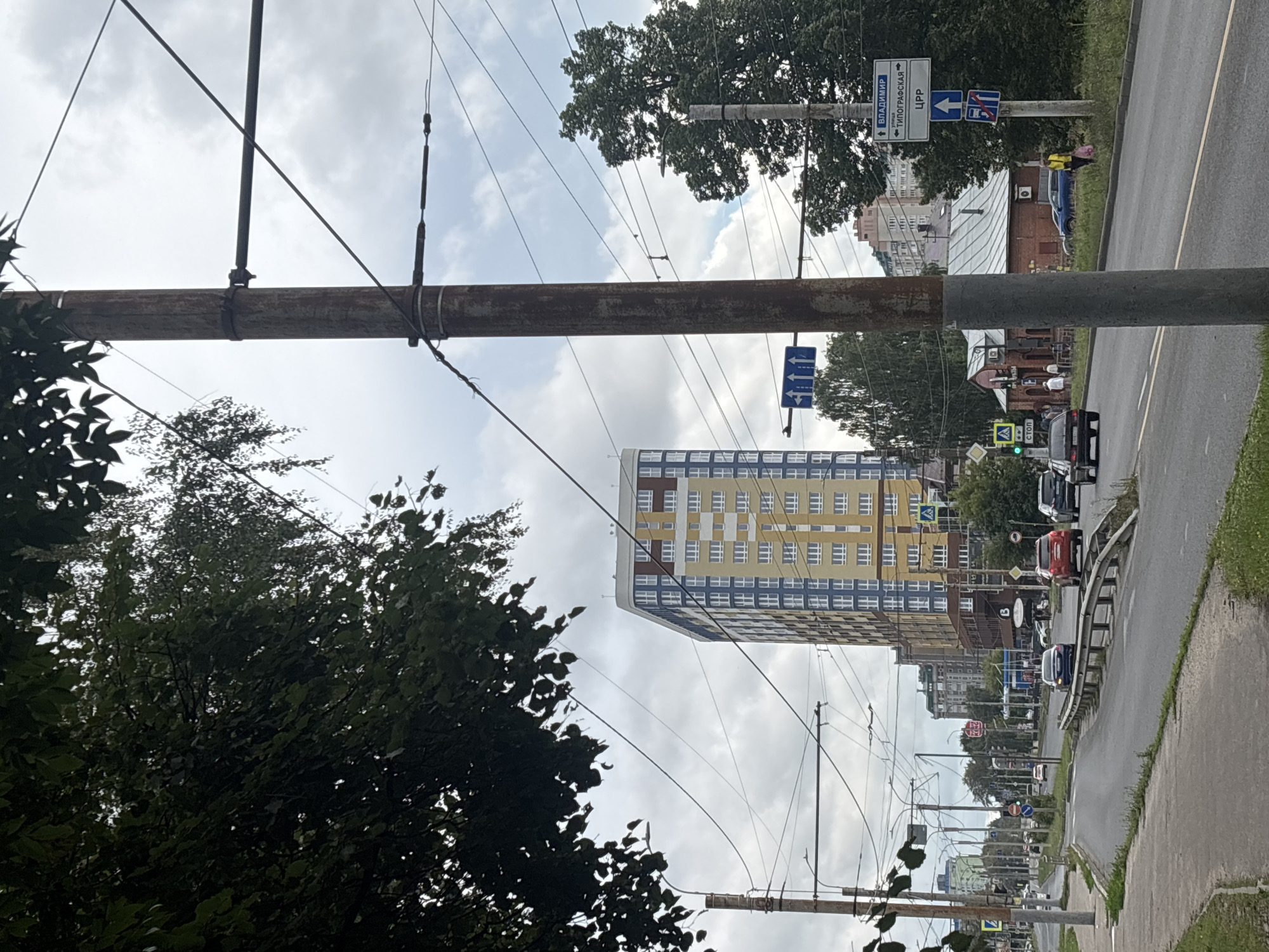 The image shows a street scene in Ivanovo, Russia, on a cloudy day. The street is lined with trees and buildings, and there are several cars on the road. In the foreground, a large utility pole with wires runs vertically. A tall, multi-story apartment building with yellow and blue facade is visible in the mid-ground. Traffic signs are present, including one indicating directions to "VLADIMIR" and "TIPOGRAFSKAYA ULITSA." Another sign shows a street closure symbol. Several pedestrians are walking on the sidewalk. The overall impression is of a typical urban street with active traffic and pedestrian activity under an overcast sky.