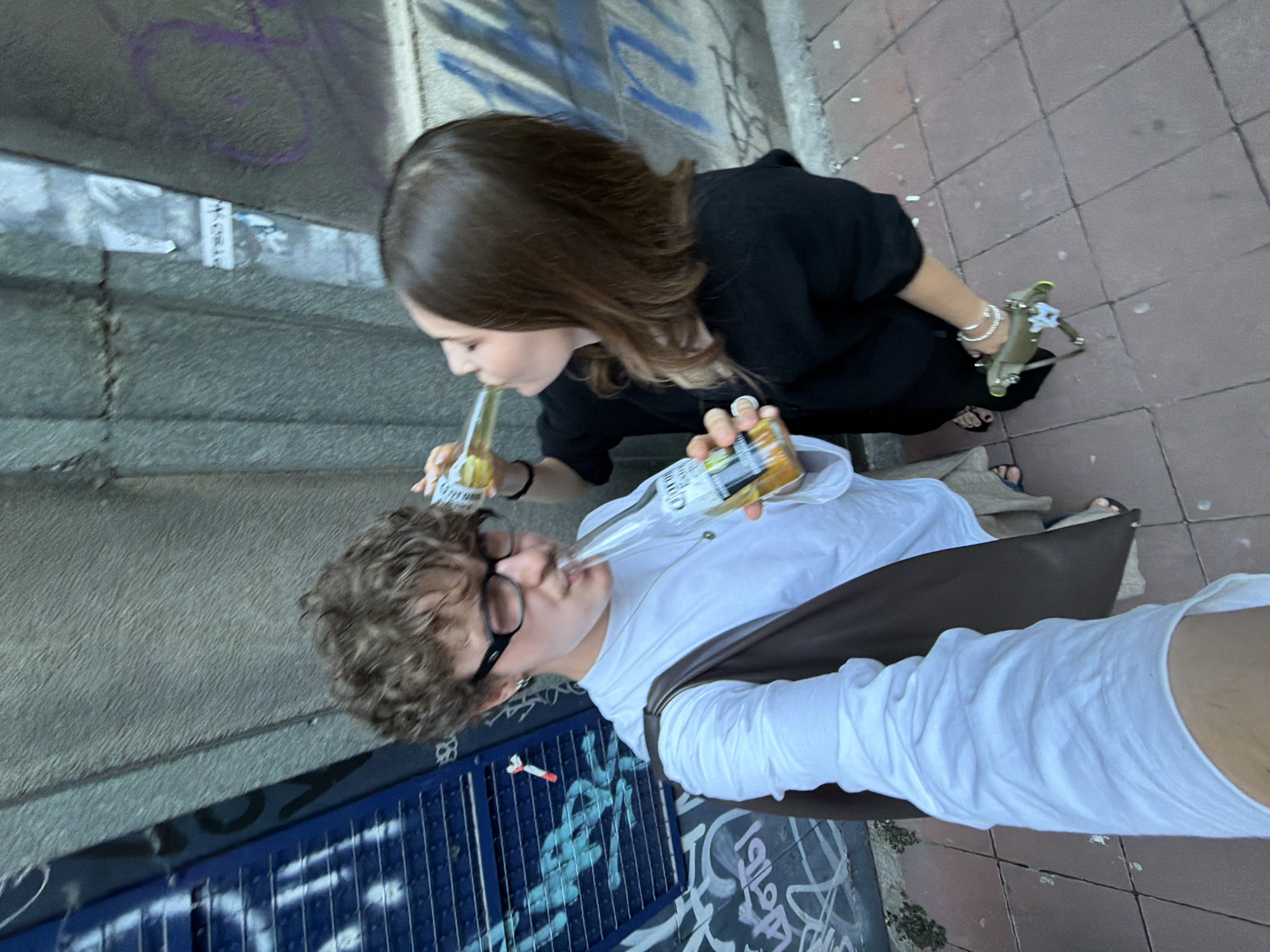 Two individuals, a male and a female, are pictured outdoors in what appears to be a city alleyway or underpass in Belgrade, Serbia. The male, on the left, has curly brown hair, is wearing glasses, a white long-sleeved shirt, and light-colored shorts. He is holding a bottle of Corona beer and drinking from it through a straw. He is also carrying a brown bag over his shoulder. The female, on the right, has long brown hair and is wearing a black top. She is also holding a bottle of Corona beer and drinking from it directly. The background consists of a rough, textured concrete wall covered in graffiti. The ground is paved with reddish-brown tiles. The lighting suggests it is daytime.