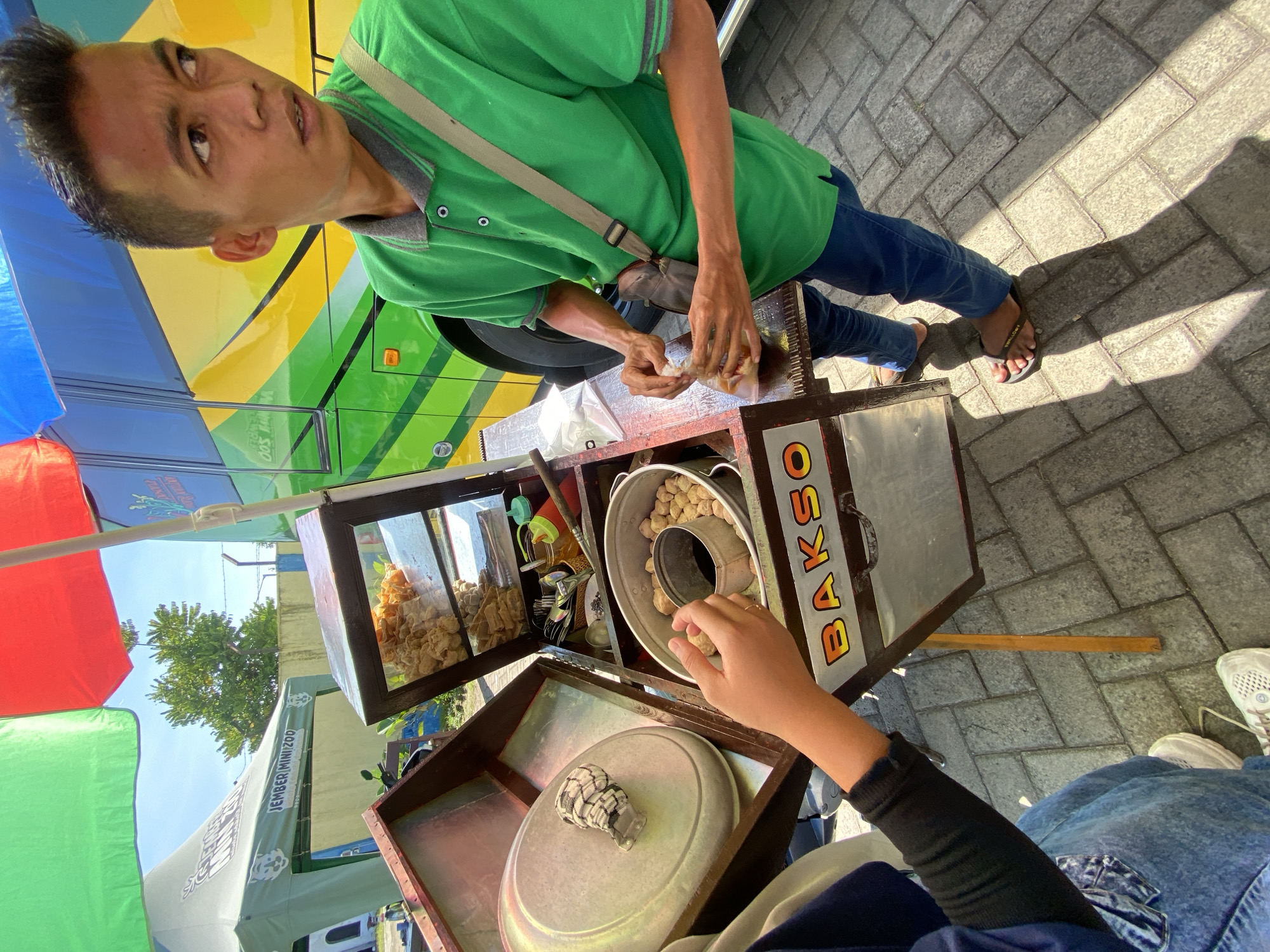 A street vendor in Mangli, Indonesia prepares a dish of Bakso. The vendor, a male with short dark hair and wearing a green polo shirt and blue jeans, stands behind his mobile food cart. His cart is made of metal with a wooden frame, and has "BAKSO" painted on its side. The cart contains a pot of meatballs, a display case of fried snacks, and other cooking utensils. A customer in blue jeans and white shoes watches as the vendor prepares their food. The ground is paved with gray bricks, and a colorful tent and a bus are visible in the background.