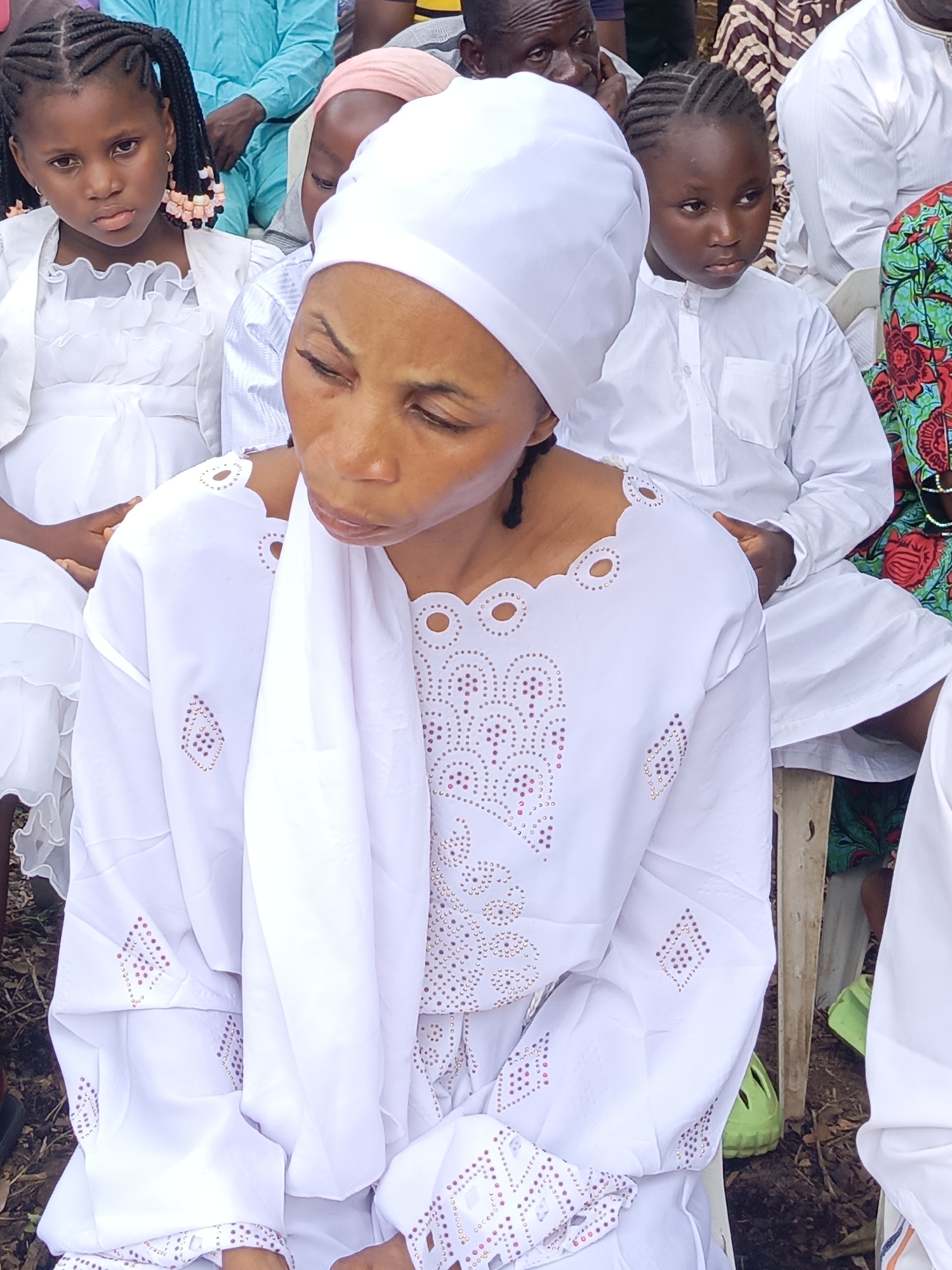 This image depicts an outdoor gathering in Andaha, Nigeria, with several individuals, predominantly women and children, dressed in white attire. The central figure is a woman with a dark complexion, seen in a three-quarter profile, wearing a white headscarf and a white long-sleeved top. Her top features intricate pink and red studded patterns, arranged in geometric shapes, and a scalloped neckline with circular cutouts. Her head is bowed, and her eyes are cast downwards, suggesting a solemn, contemplative, or prayerful mood.
To the left of the central woman, a young girl, approximately 6-8 years old, looks directly at the viewer with a serious expression. She has dark braided hair adorned with pink and light brown beads and is dressed in a white ruffled dress and a matching white jacket. On the right side, a young child, possibly a boy, with braided hair and a white long-sleeved shirt with a small collar, looks off to the right with a pensive gaze.
The background features blurred figures of other attendees, some also in white, and others in patterned garments, suggesting a larger congregation. Glimpses of a man's face are visible in the upper left background, and another man in a light blue traditional garment is further behind. The setting is outdoors, indicated by the dirt ground and scattered dry leaves. Partial views of simple chairs suggest that the people are seated. A bright green plastic sandal or shoe is partially visible on the ground in the lower right. The natural, bright lighting indicates it is daytime. The overall mood appears respectful and serious, characteristic of a religious ceremony or a significant community event. No text is visible in the image.