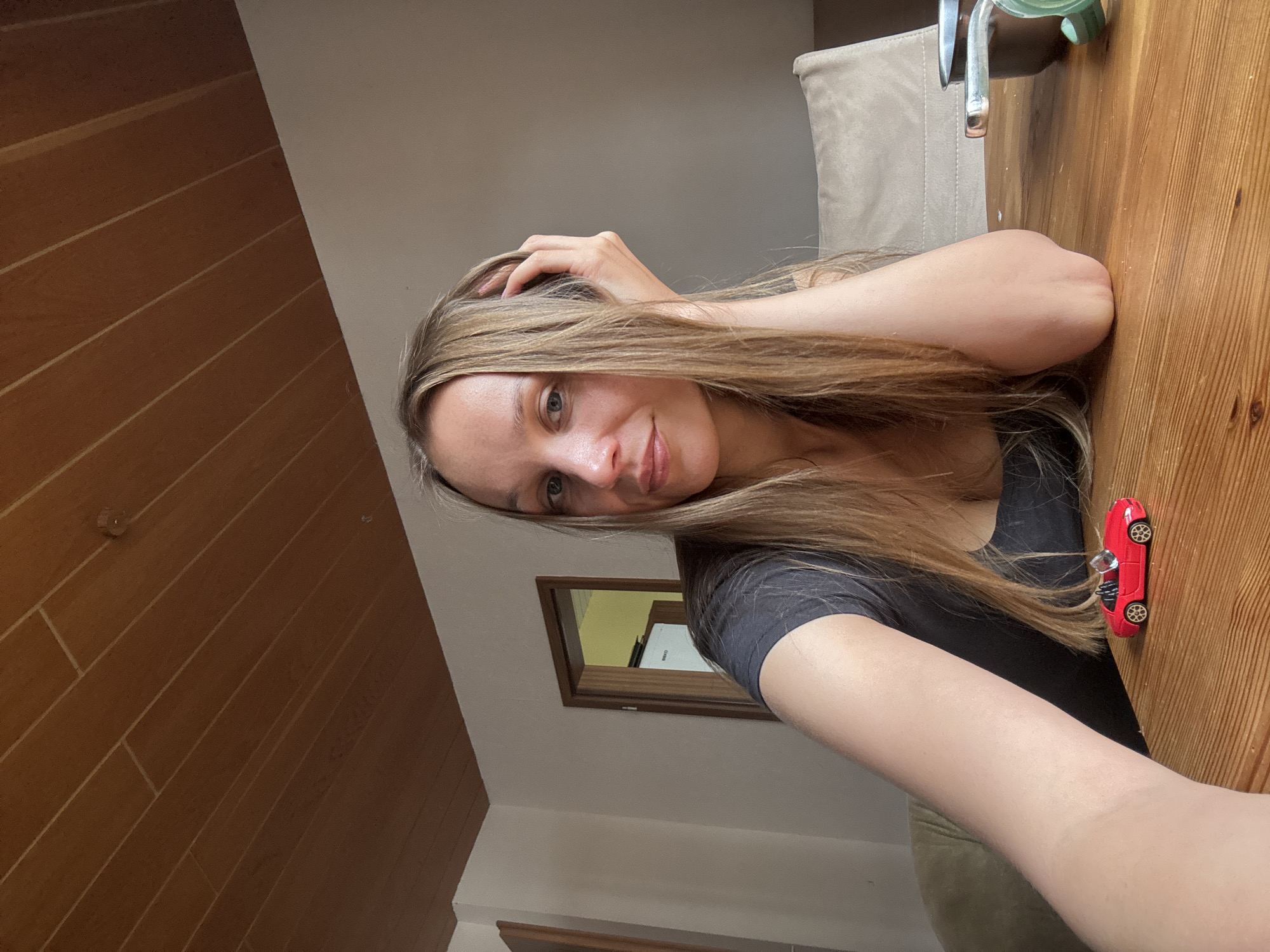 A woman with long blonde hair is sitting at a wooden table, with her left arm resting on the table and her right hand touching her hair. She is looking at the camera with a slight smile. On the table in front of her is a small red toy car. In the background, there is a door, a wall, and a ceiling with wooden panels. The scene appears to be indoors, possibly in a home. The image does not contain any information about the city of Überherrn, Germany.