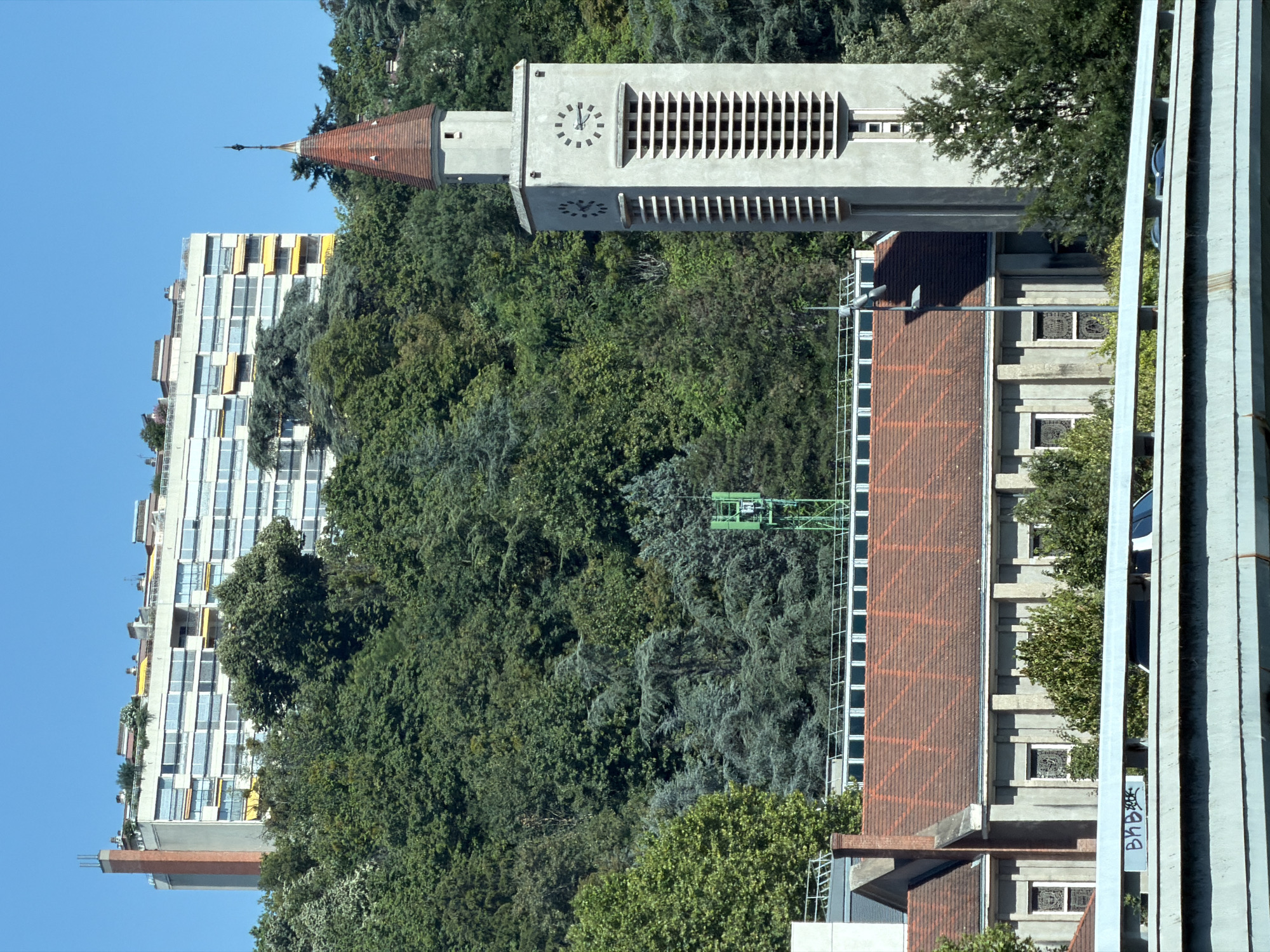 The image shows a view of the city of Lyon, France, dominated by a large, modern apartment building situated on a hillside. In the foreground, a church with a red tile roof and stained glass windows is visible. To the right of the church is a tall, slender bell tower with a conical red roof, featuring a clock face and decorative grilles. The hillside is densely covered with various trees, creating a green backdrop. A green cell tower is also partially visible among the trees. In the far distance, a bridge spans across the bottom of the frame, with graffiti visible on its railing. The sky is clear and blue, indicating a sunny day. No people are clearly identifiable in the image.
