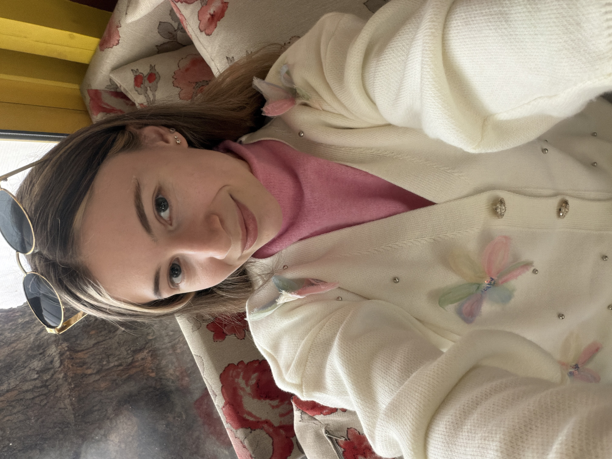 A young woman is taking a selfie. She has fair skin, brown hair with blonde highlights, and is wearing sunglasses on her head, a pink top, and a white cardigan with floral accents. She appears to be indoors, with a patterned fabric behind her. Outside the window, one can see the city of Plovdiv, Bulgaria.