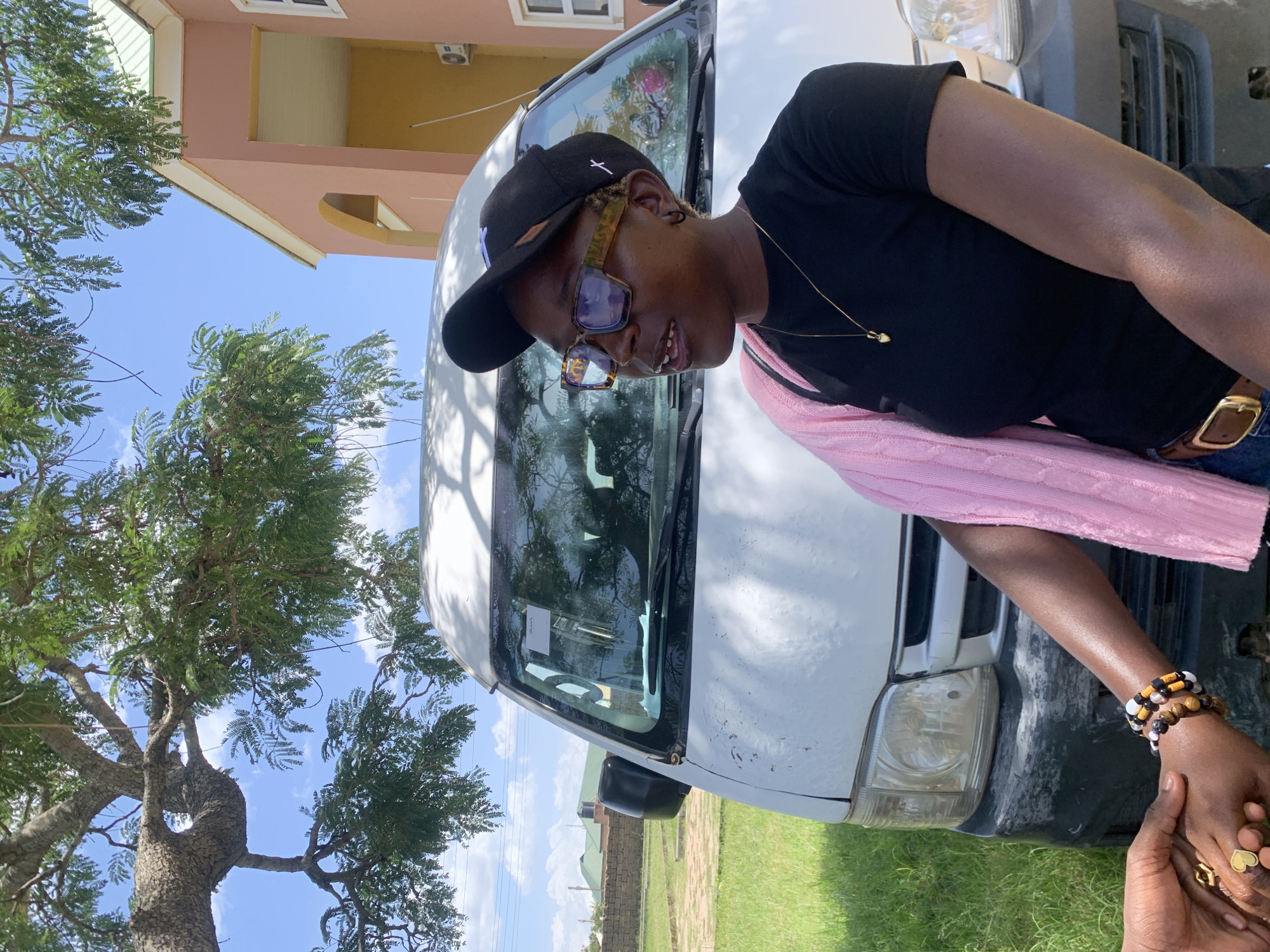 The image depicts two individuals, seemingly holding hands, in an outdoor setting in Gwafan, Nigeria. The primary subject is a person wearing a black t-shirt, a black baseball cap, and distinctive glasses with patterned frames. They are leaning against a white van, with their right arm resting on the vehicle. A pink knitted garment is draped over their left shoulder and across their lap. They are also wearing several bracelets on their right wrist, and rings on their left hand. The second person's hand is visible in the foreground, also wearing rings.
The scene appears to be during the daytime, under a clear blue sky with some scattered clouds. A large tree with green foliage is prominently featured on the left side of the image, casting shadows. In the background, a building with pink and yellow sections is visible, suggesting a residential or institutional structure. The white van appears to be parked on a grassy area. The overall mood seems casual and relaxed, with the subjects appearing comfortable with each other. There is no visible text in the image.