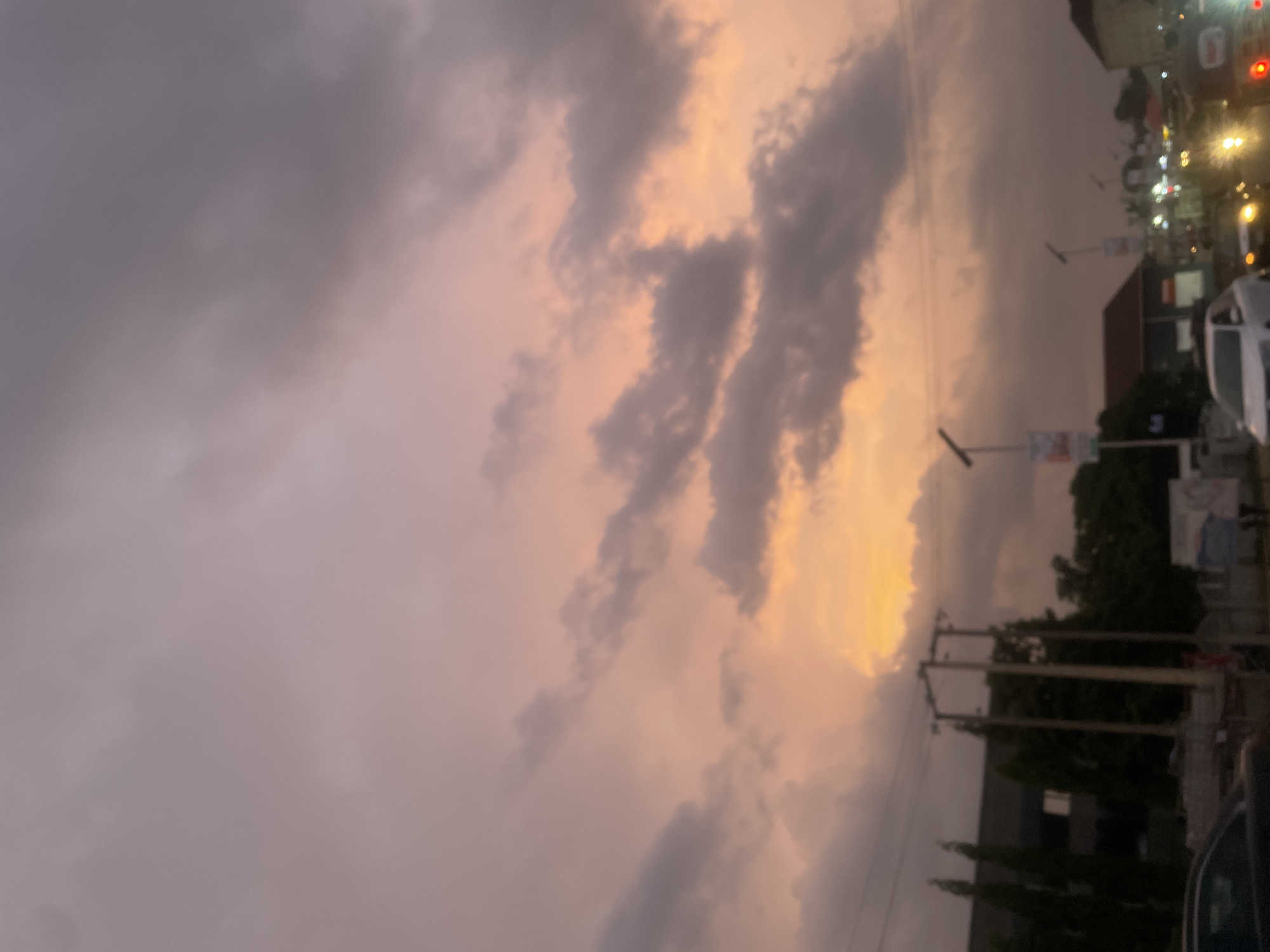 The image shows a cloudy sky with a pink and orange glow, possibly at sunset or sunrise, over a street scene in Abuja, Nigeria. Utility poles and wires are visible along the side of the road. There are trees and buildings along the street, as well as cars. Streetlights are on, suggesting it is either dusk or night.