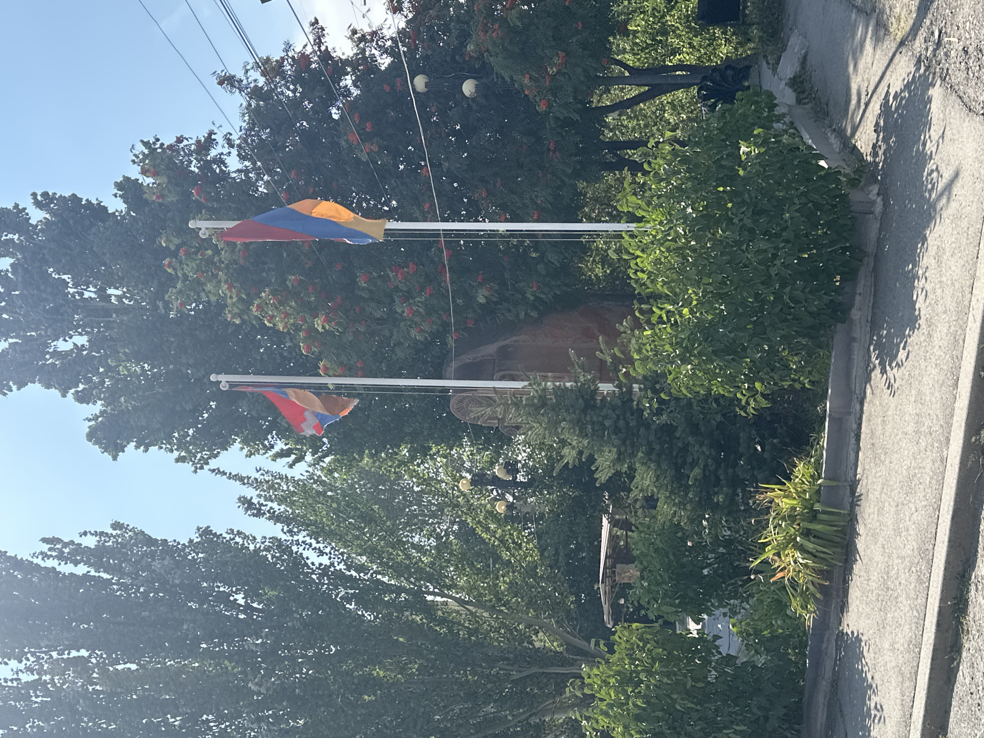 The image depicts a scene in Gyumri, Armenia, under a clear blue sky. Two Armenian flags are prominently displayed on white flagpoles in the foreground. The flags are red, blue, and orange. Behind the flags, a dense cluster of trees with red berries is visible, suggesting it might be late summer or early autumn. To the left of the flags, tall trees with dark green foliage create a lush backdrop. A street light with multiple globes can be seen amidst the greenery. In the lower part of the image, a paved sidewalk and road are present, with shadows cast by the trees and foliage. The overall impression is a bright, outdoor setting in an urban or park-like area of Gyumri.