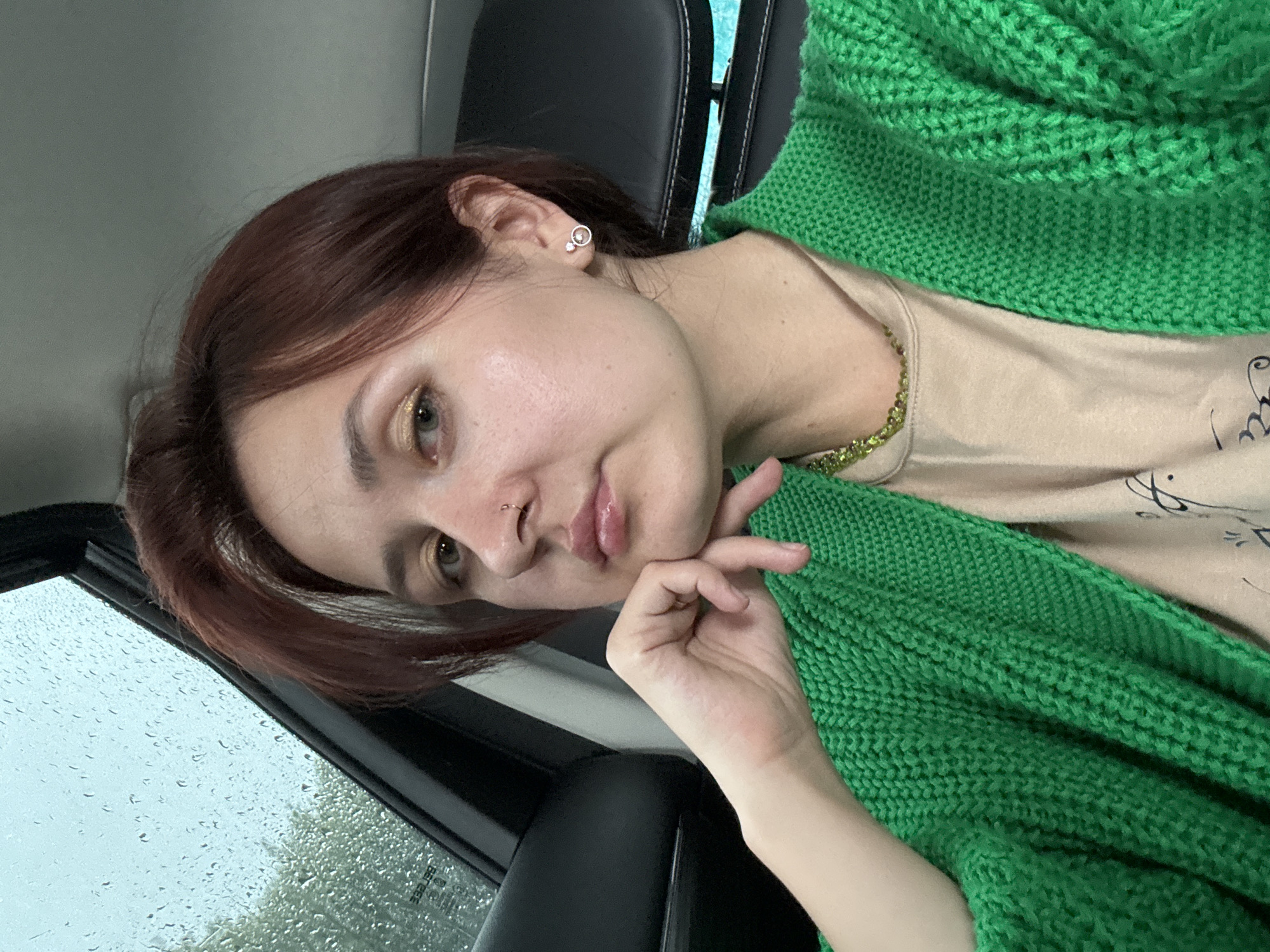 A young woman with short, reddish-brown hair and light skin is pictured from the chest up, inside a vehicle. She is wearing a bright green knitted cardigan over a tan t-shirt and a green beaded necklace. Her left hand is resting on her chin, and she has a nose ring and small hoop earrings. The background shows a car window with raindrops, suggesting an outdoor environment, possibly during inclement weather. The scene is captured in a close-up, selfie-style shot.
