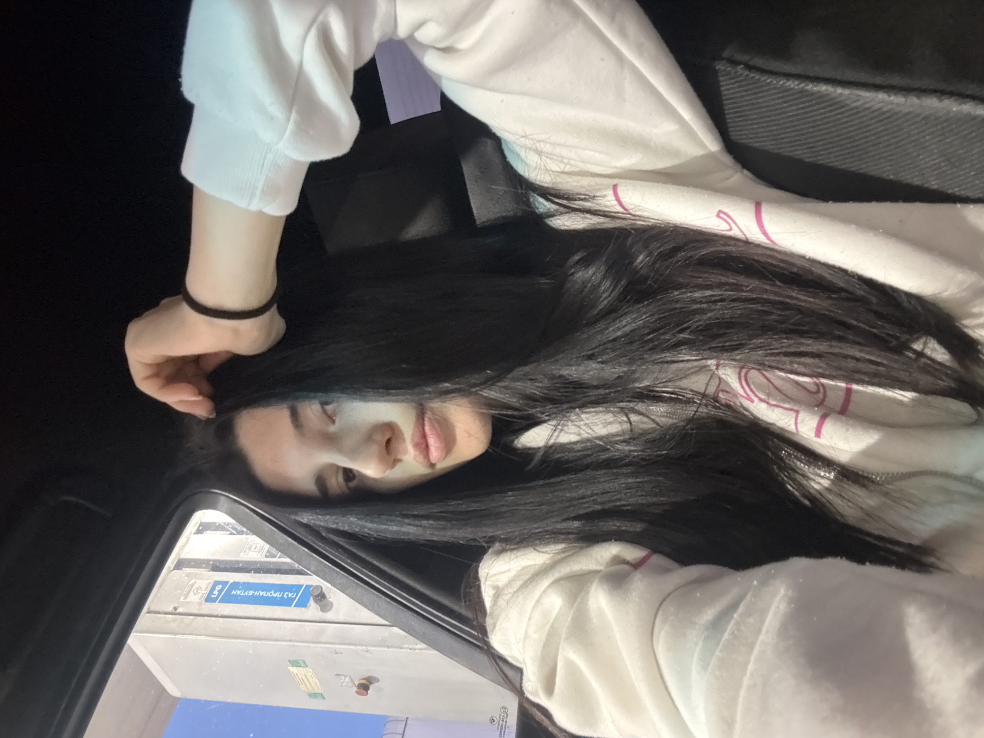 A woman with long, dark hair rests her head on her hand inside a car parked at a gas station. She is wearing a white hoodie with pink accents. The gas pump visible through the car window has Cyrillic text, reading "ГАЗ ПРОПАН-БУТАН LPG", which indicates that it dispenses propane-butane gas. This gas station is likely located in Sofia, Bulgaria due to the Cyrillic text.