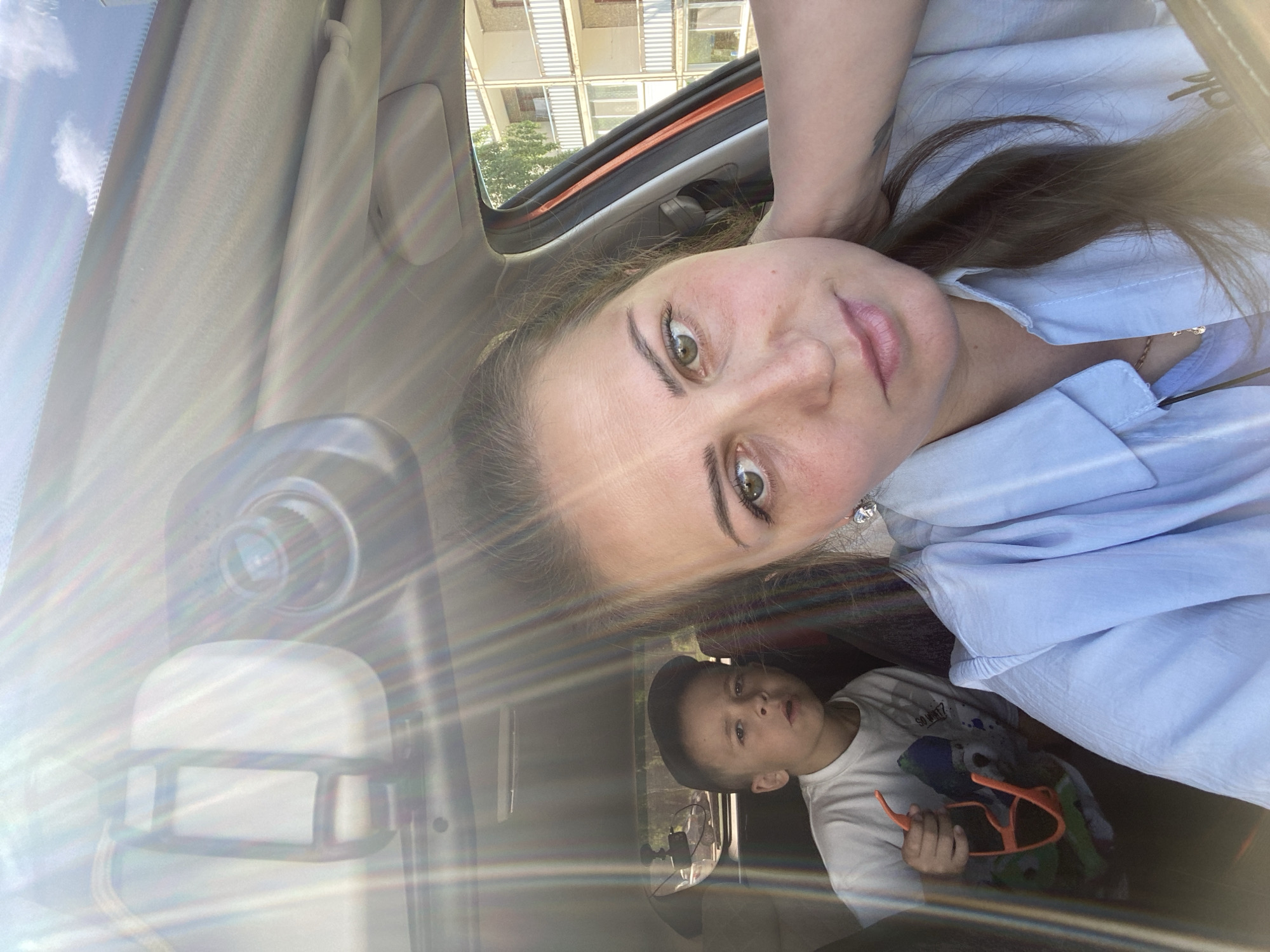 In the image, a woman and a young boy are inside a car. The woman is in the foreground, taking a selfie. She has long brown hair and is wearing a light blue collared shirt. A young boy is visible in the background, sitting in the back seat, wearing a white t-shirt and a dark baseball cap. He is holding a pair of orange sunglasses. The interior of the car is visible, including the rearview mirror and part of the dashboard. Sunlight streams into the car, creating lens flares. Outside the car, a multi-story building with balconies can be seen, suggesting an urban or suburban setting. This image could have been taken in the city of Марковский, Russia.