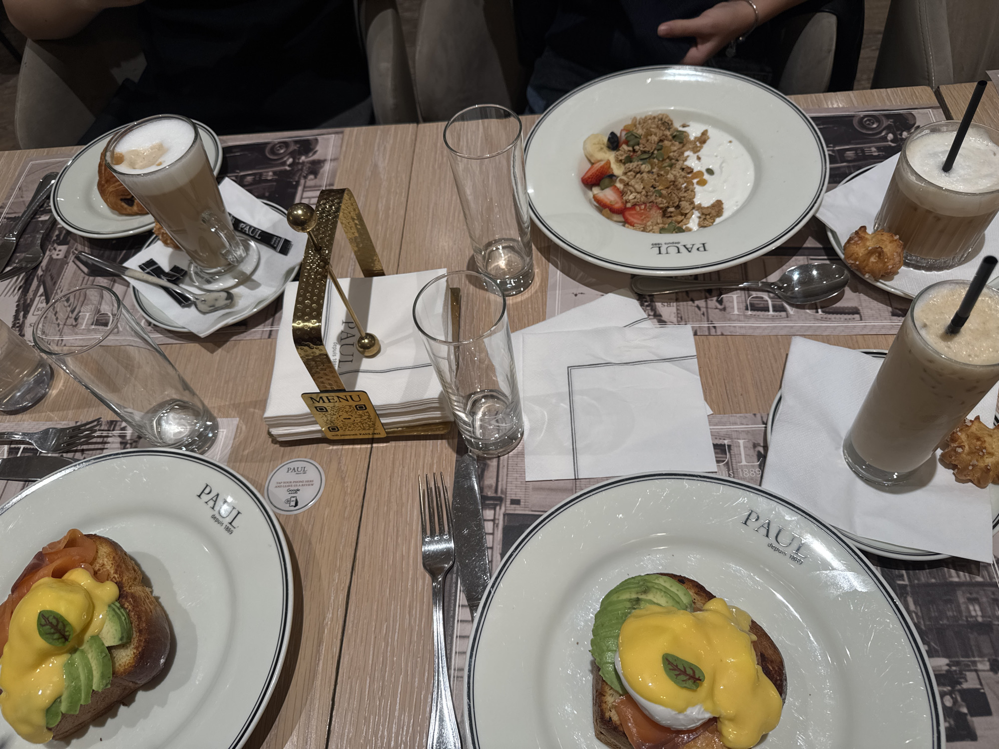 An overhead shot captures a dining table set for breakfast or brunch at a PAUL cafe in Yerevan, Armenia. Two individuals are present, indicated by visible arms and hands. The wooden table is covered with paper placemats featuring a black and white cityscape design.
Various dishes and drinks are spread across the table. On the left, a tall glass of latte with foam sits next to a croissant. Below it, a plate holds a dish consisting of brioche or toast topped with smoked salmon, avocado slices, a poached egg, and hollandaise sauce, garnished with a small green leaf. Another identical dish is visible on the right side of the table.
In the upper right, a white bowl contains yogurt topped with granola, strawberries, banana slices, pumpkin seeds, and dried fruit. Beside it, two iced coffee or latte drinks with black straws are served in glasses, accompanied by small, star-shaped biscuits. Several empty water glasses and cutlery (forks, knives, spoons) are also present. A golden menu holder, also serving as a napkin dispenser, stands prominently in the center.
Visible text includes the brand name "PAUL" and "depuis 1889" (since 1889) on the white plates and placemats. A small golden card reads "MENU" with QR codes and "IG: @paul_armenia". A round sticker invites customers to "TAP YOUR PHONE HERE AND LEAVE US A REVIEW" with the Google logo. The setting suggests a relaxed and enjoyable meal, likely taking place in the morning or early afternoon.