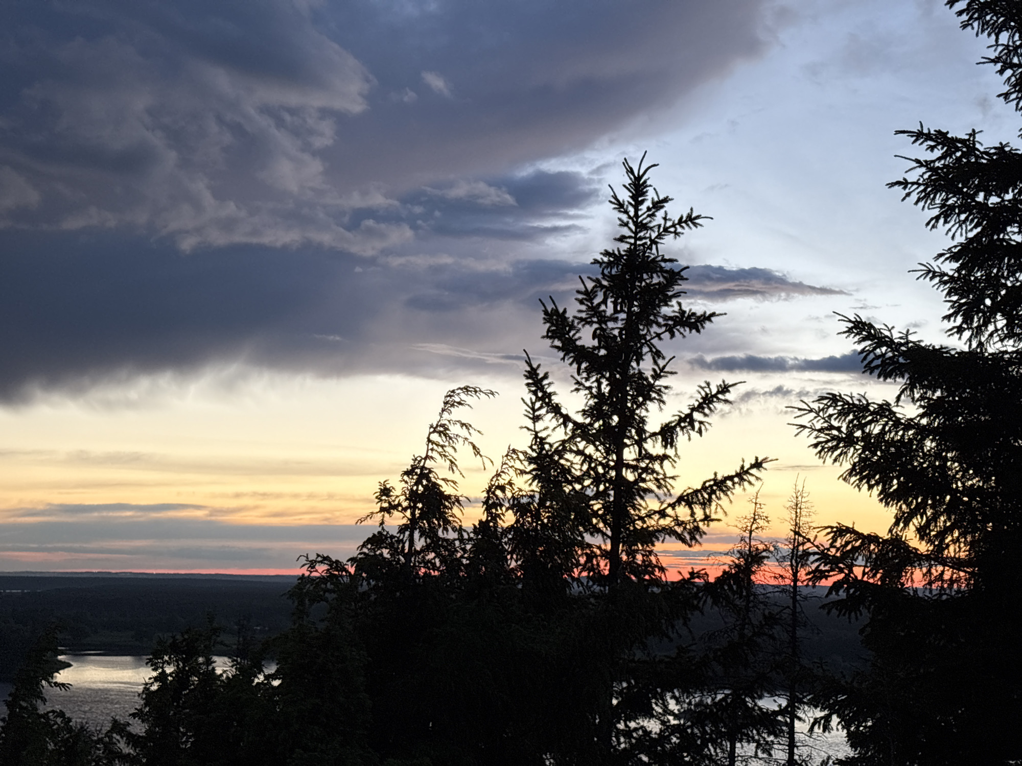 The image shows a landscape view at sunset or sunrise in Ольховка, Russia. The foreground is dominated by silhouetted trees, primarily evergreens, obscuring the full view of the scene. In the middle ground, there is a body of water (possibly a river or lake) and a stretch of land with dense vegetation. The background displays a colorful sky with a gradient of oranges, pinks, and purples near the horizon transitioning to blues and grays higher up, with prominent cloud formations, particularly darker clouds covering the upper left portion of the frame. The overall scene evokes a sense of serenity and natural beauty.