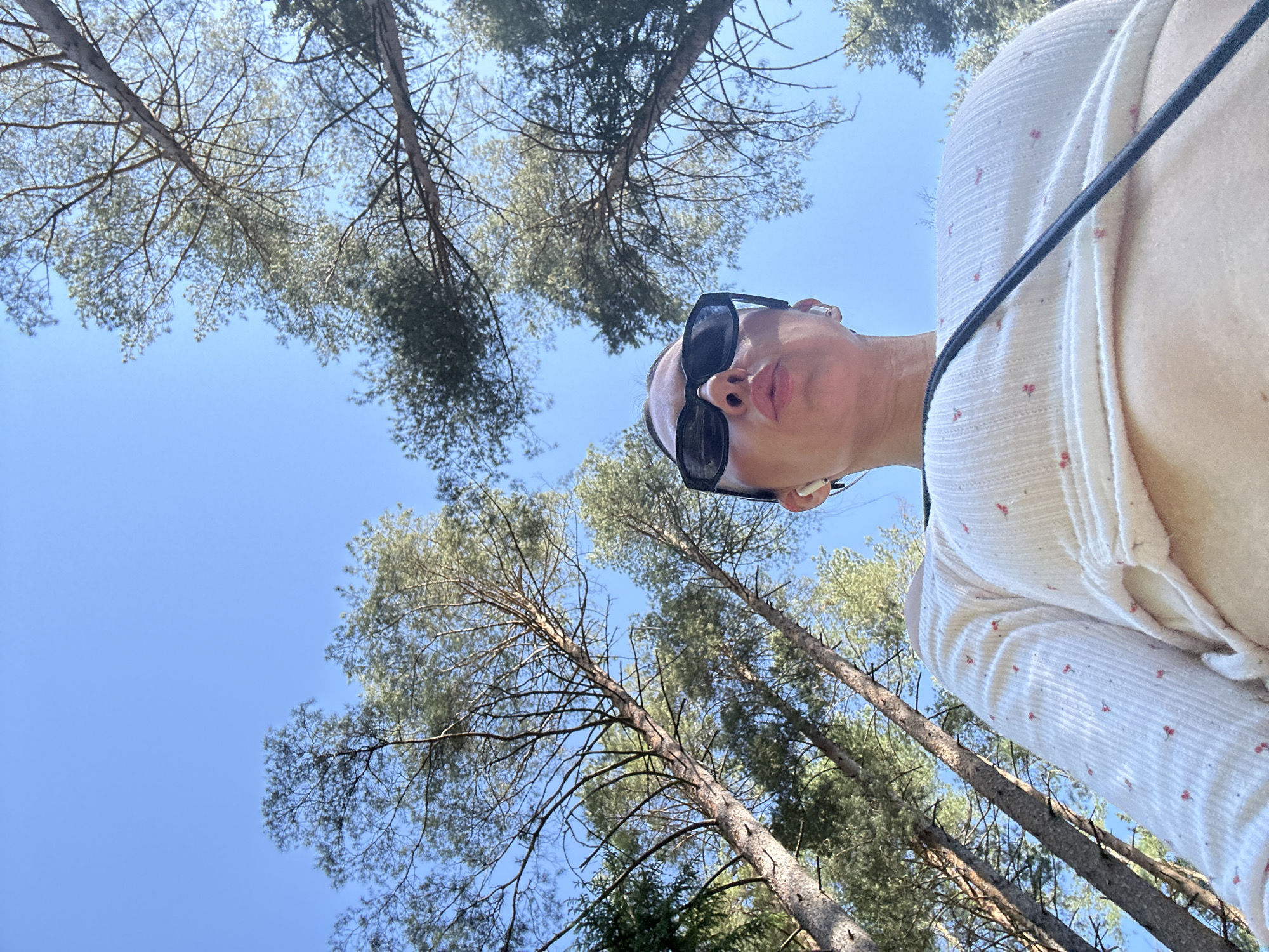 The scene is an upward-angled selfie taken outdoors, likely in a forested area in Votkinsk, Russia. A person with sunglasses and an earpiece is in the foreground. Overhead are tree branches against a bright blue sky. The person is wearing a light-colored shirt with small red patterns. A black strap is visible over the person's shoulder. The environment includes tall pine trees.