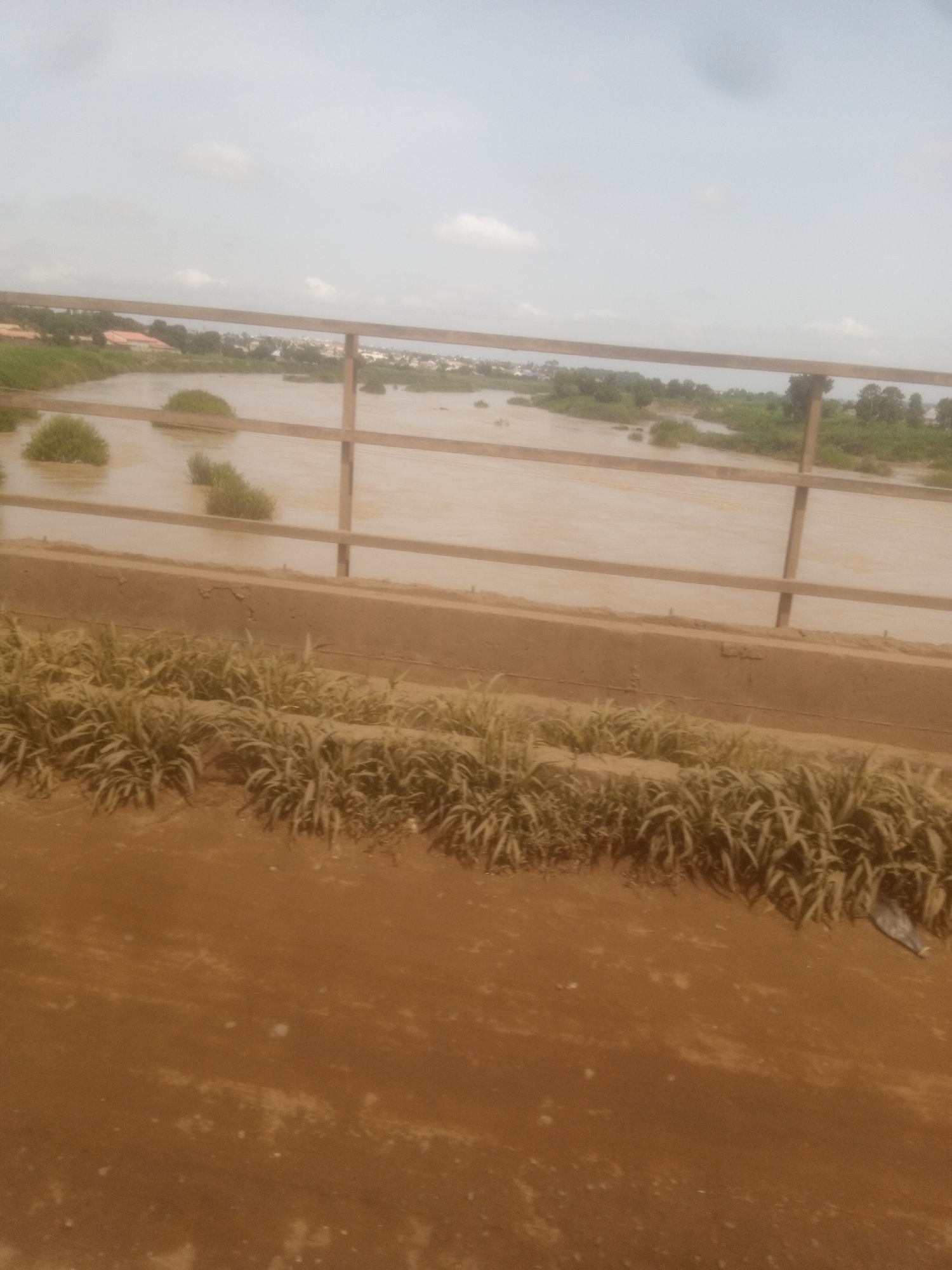 A wide, brown river flows through a landscape with green vegetation and some scattered buildings on the far bank. In the foreground, a metal railing with vertical posts runs horizontally across the frame, separating the viewer from the river. The railing is supported by a concrete structure. Dry, light brown soil and patches of tall, dry grass are visible in the immediate foreground. The sky is overcast with pale clouds. The scene appears to be a view from a bridge in Nassarawa, Nigeria, overlooking the river and its surroundings.