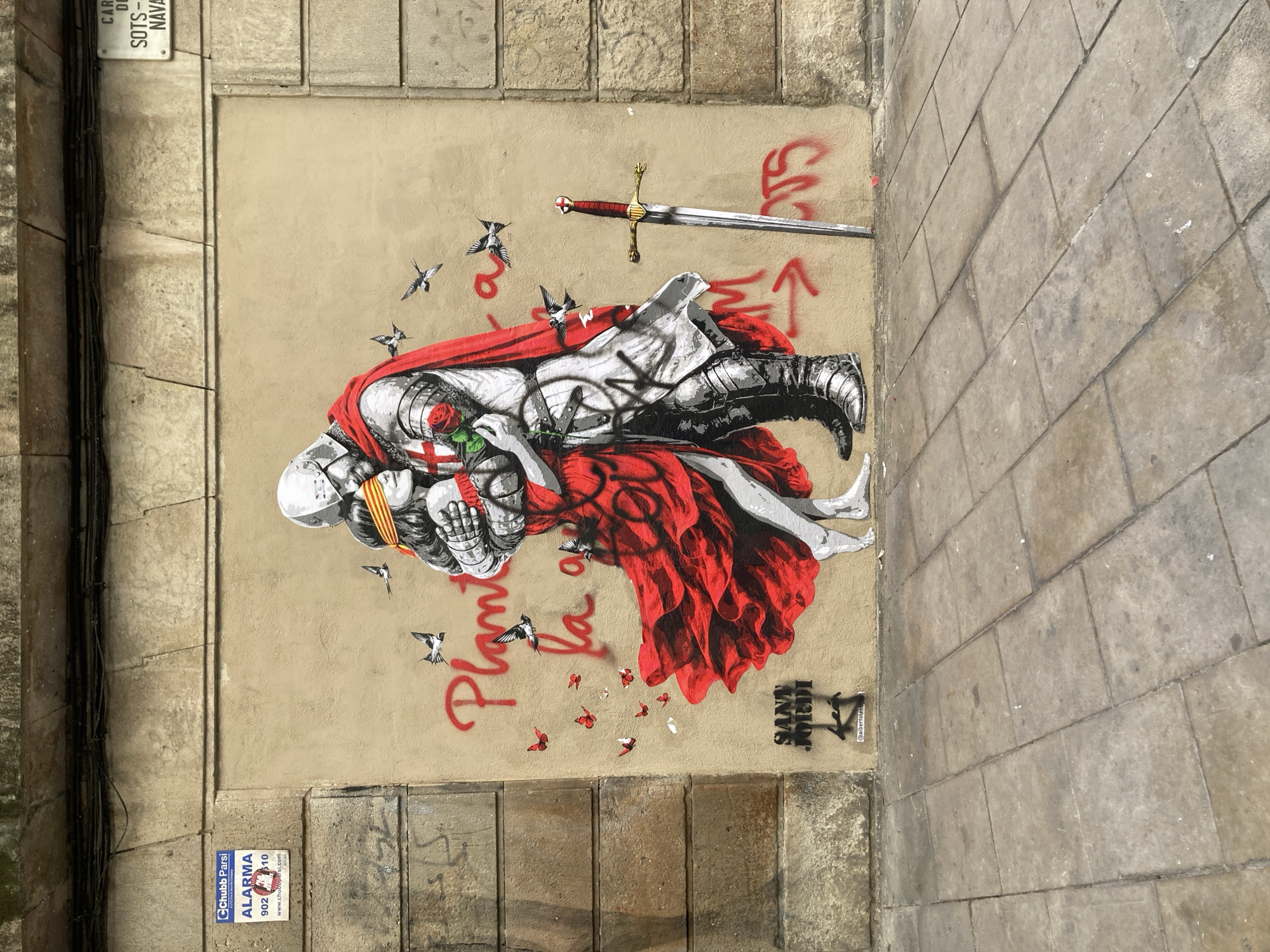 The image shows street art on a textured wall in Barcelona, Spain. The artwork depicts a knight in armor, holding a rose, with a red cape or cloth draped over him. He is lying next to a figure in white with bare feet. A sword lies horizontally above them. Several swallows fly around them. Red text reads "Planto la," and below that, "Sant Jordi," along with the artist's tag. The wall also has a sign with "CARL DL SOTS- NAVA" and an alarm sign that says "Chubb Parsi ALARMA 902...". The wall is bordered by stone. The photo is taken at a slight angle.