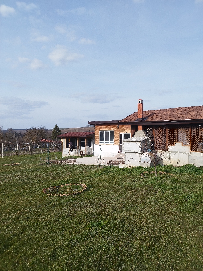 The scene, located in Odrintsi, Bulgaria, is an outdoor area under a partially cloudy blue sky. The foreground is dominated by green, uneven grass, with a circular arrangement of small stones or debris in the lower-left quadrant. Two single-story buildings are present in the mid-ground. The primary structure, positioned on the right, features a brown shingle roof with a prominent brick chimney. Its walls consist of light-colored lower sections and reddish-brown upper paneling, complemented by multiple white-framed windows and a screened porch area with dark wooden lattice on its right side. Directly in front of this building is a light gray concrete block outdoor cooking structure, flanked by small wooden fences. To the left and slightly behind, a secondary, smaller single-story building with a reddish-brown roof and a covered entrance is visible. One individual, dressed in dark clothing, is standing near the entrance of this secondary building. The background displays bare deciduous trees, a single evergreen tree, and distant rolling terrain.