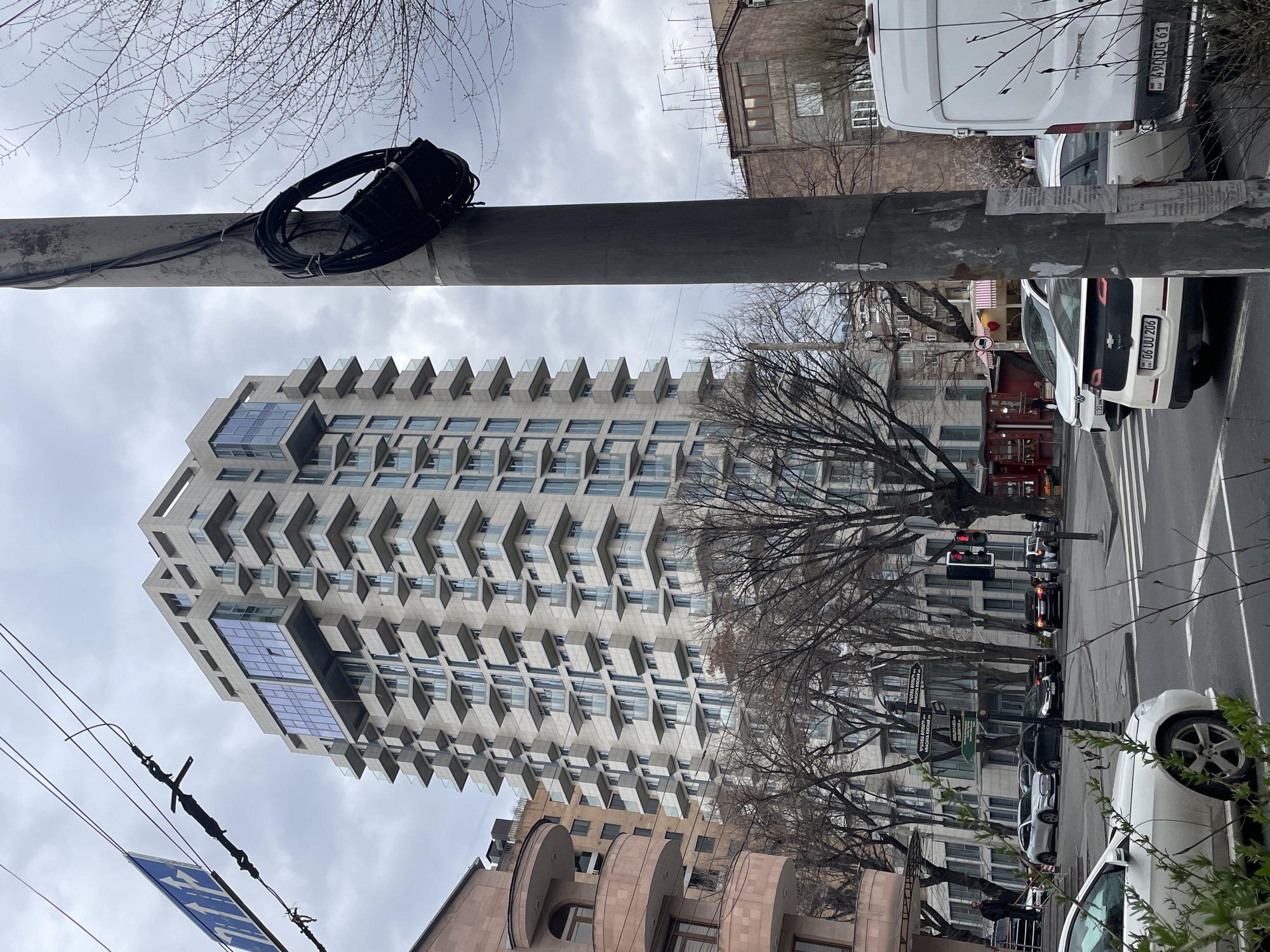 The image captures a street scene in Yerevan, Armenia, featuring a modern, tall building with projecting balconies, situated among older buildings. A utility pole with attached wires and equipment runs vertically through the frame, partially obscuring the view. A line of parked and moving vehicles is visible along the street, including cars and a white van. Bare trees line the street, indicating a colder season. Street signs are mounted on poles along the street. The sky is overcast.