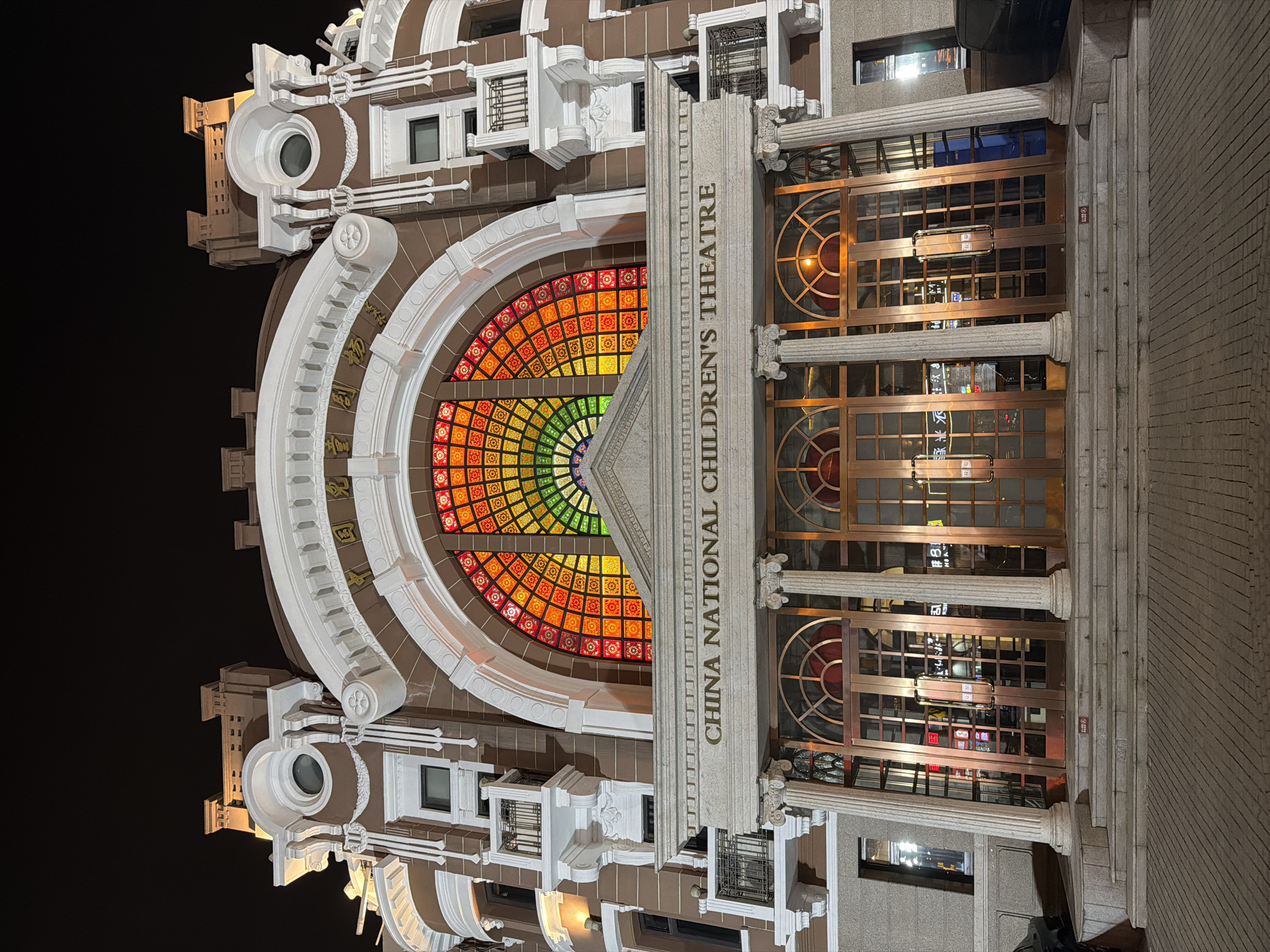 The image shows the China National Children's Theatre, located in the Dongcheng District of China. The building features a large, semi-circular stained glass window with a rainbow color pattern above the entrance. The entrance is flanked by decorative columns and has several sets of double doors. The building's facade is detailed with white architectural ornamentation on a brown base. The image appears to be taken at night, as the background is dark and the building is illuminated.