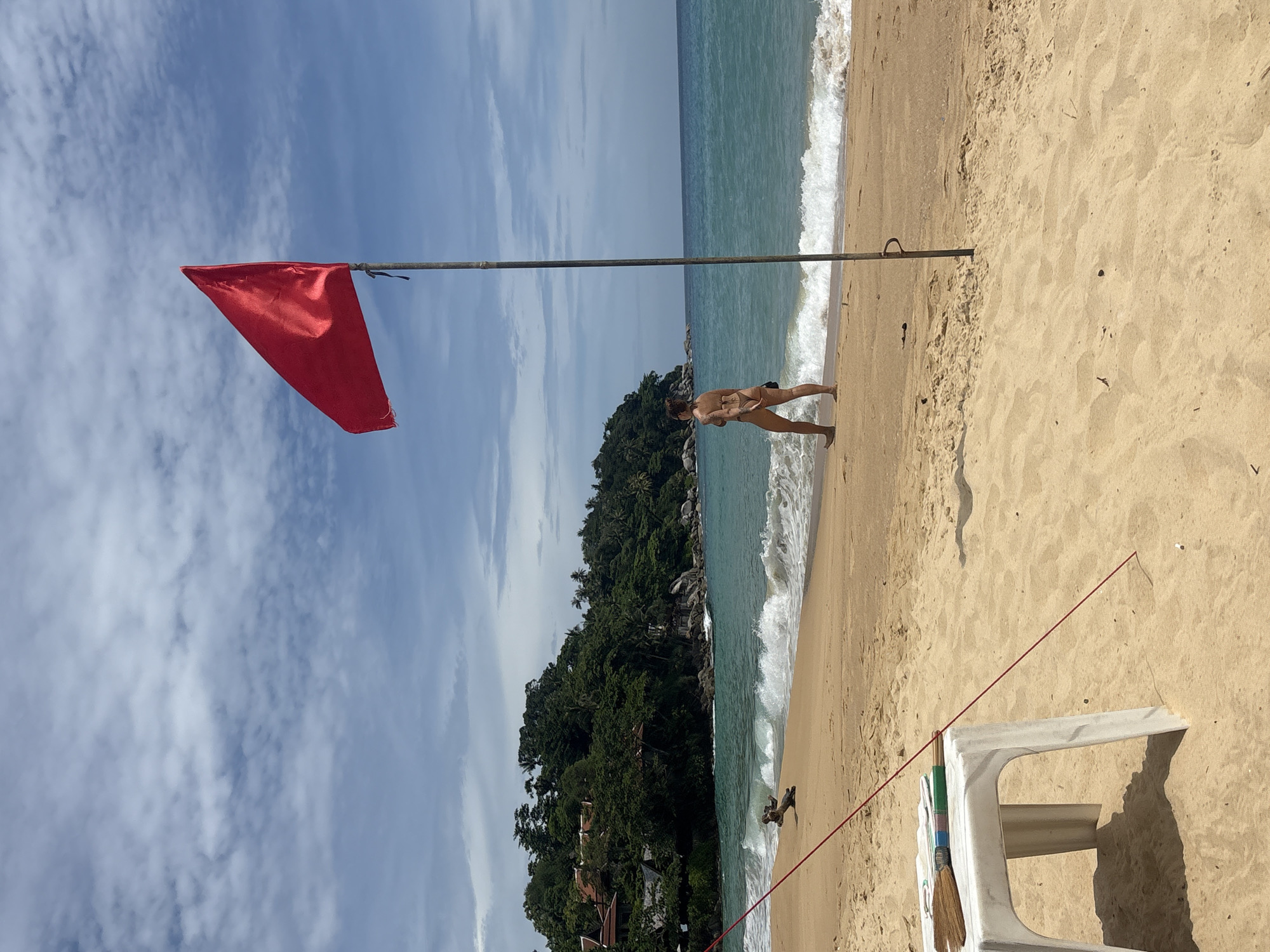 The image captures a serene beach scene in Karon, Thailand. The foreground features a sandy beach with a weathered white plastic chair and a red flag planted on a pole, which is tilted. The flag is red and billows slightly in the breeze. The sea, with gentle waves, is visible on the left side of the image, and the water is a clear turquoise.
In the middle ground, a woman stands on the wet sand near the waterline. She is wearing a bikini and appears to be looking out at the ocean. Behind her, a lush green, tree-covered hill rises from the beach, dotted with some buildings, suggesting a tropical environment. The sky is a bright blue with scattered white clouds, indicating fair weather and a sunny day. The time of day appears to be late morning or early afternoon, given the bright sunlight.
Further in the background, the horizon shows a clear delineation between the sea and the sky. The overall mood of the image is peaceful and relaxed, typical of a beach vacation. There is no visible text in the image.