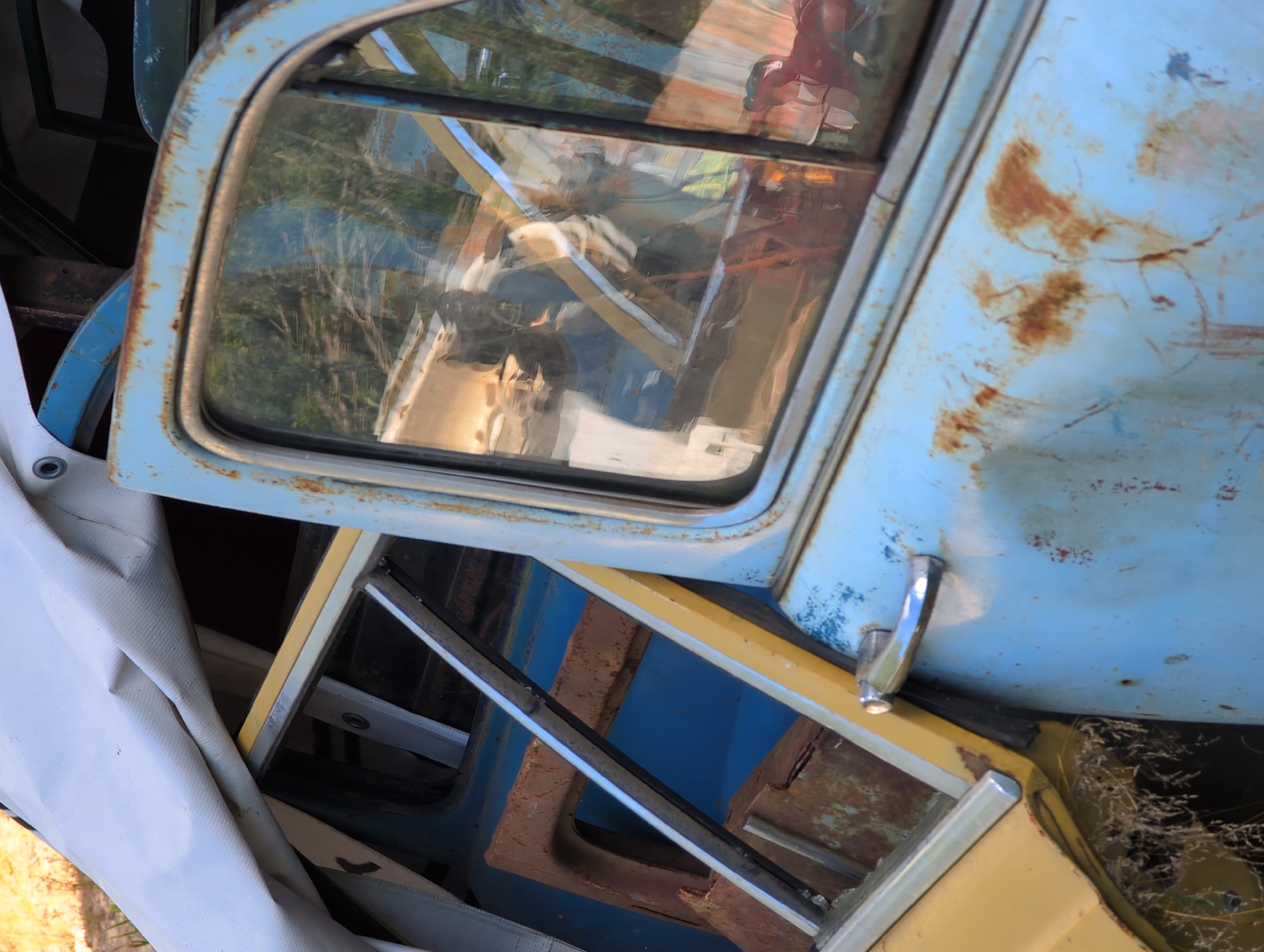 The image depicts a close-up, angled view of a derelict, light blue and yellow vehicle, heavily rusted, with a white tarp partially covering it. The vehicle's door and window are visible, with reflections of trees and foliage on the glass. Rust patches are prominent on the blue painted surface, as is a chrome door handle. The background shows dry grass and hints of other metallic objects, suggesting an outdoor storage or junkyard environment. The location is specified as Novoe Devyatkino, Russia.