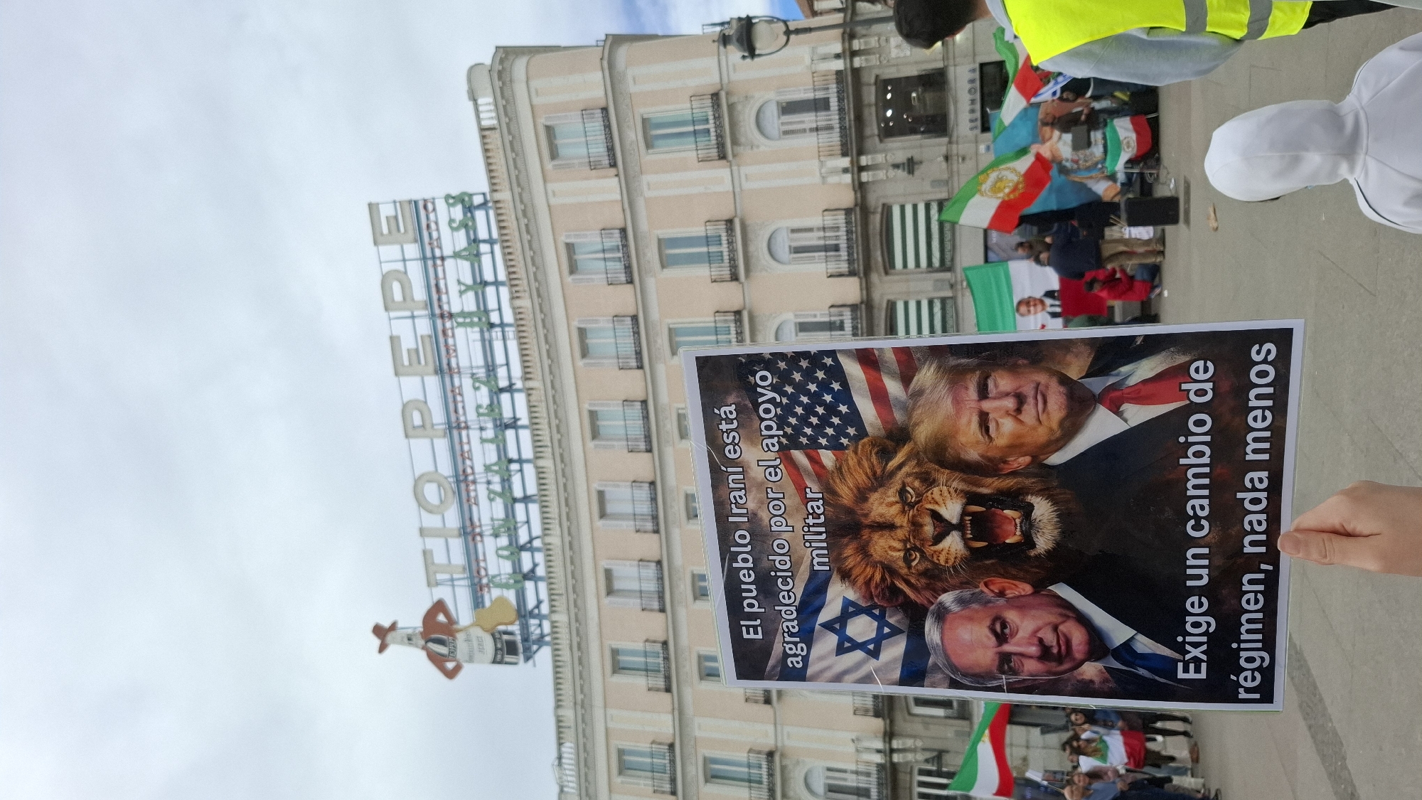 The image captures a protest scene in Madrid, Spain. In the foreground, a sign is held up displaying images of Donald Trump and Benjamin Netanyahu superimposed over an American and Israeli flag, respectively, with a lion in between. The text on the sign in Spanish reads "The Iranian people are grateful for military support. Demands a change of regime, nothing less."
In the background, people hold Iranian flags. Some individuals are wearing bright yellow safety vests. In the background is the Tio Pepe neon sign over a building in the Puerta del Sol square, a major public space in Madrid.