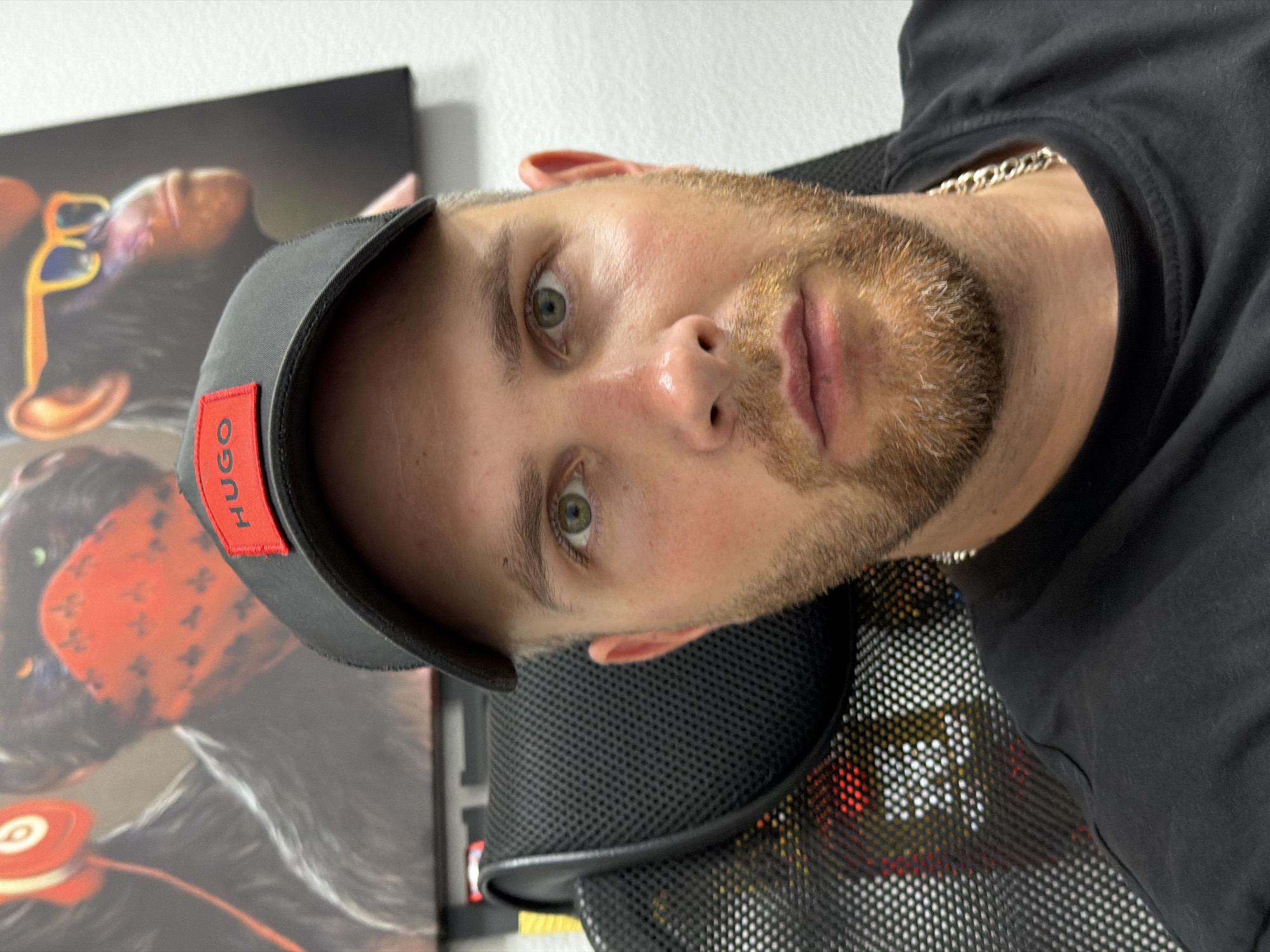 A man with a beard and a black baseball cap with a red "HUGO" logo is sitting in a black mesh chair. He is wearing a black t-shirt and a silver chain. Behind him, there are two paintings of monkeys. The painting on the left shows a monkey wearing headphones. The painting on the right shows a monkey wearing sunglasses. The man is looking at the camera. The image appears to be taken indoors, possibly in a room. There is no visible indication of the city of Chaikovsky, Russia.