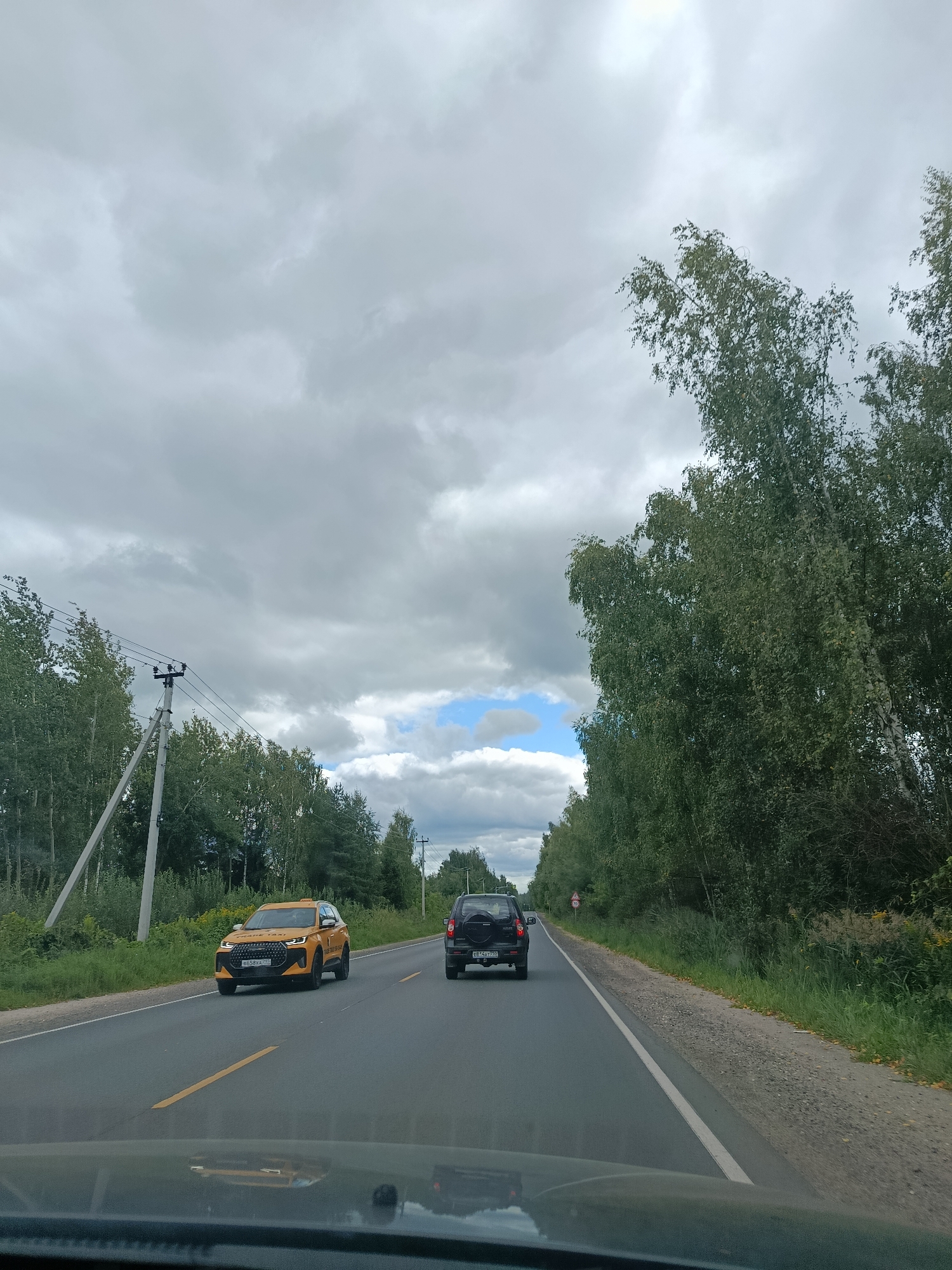 The image depicts a two-lane asphalt road with white and yellow lane markings, bordered by dense green trees on both sides. The sky is overcast with gray clouds, but a small patch of blue sky is visible. Power lines and a utility pole are present on the left side of the road. Two vehicles are driving on the road: a yellow SUV in the left lane and a black SUV in the right lane. The yellow SUV has a license plate visible. The viewpoint is from inside another vehicle, looking forward. The location is identified as 1-Go Maya, Russia.