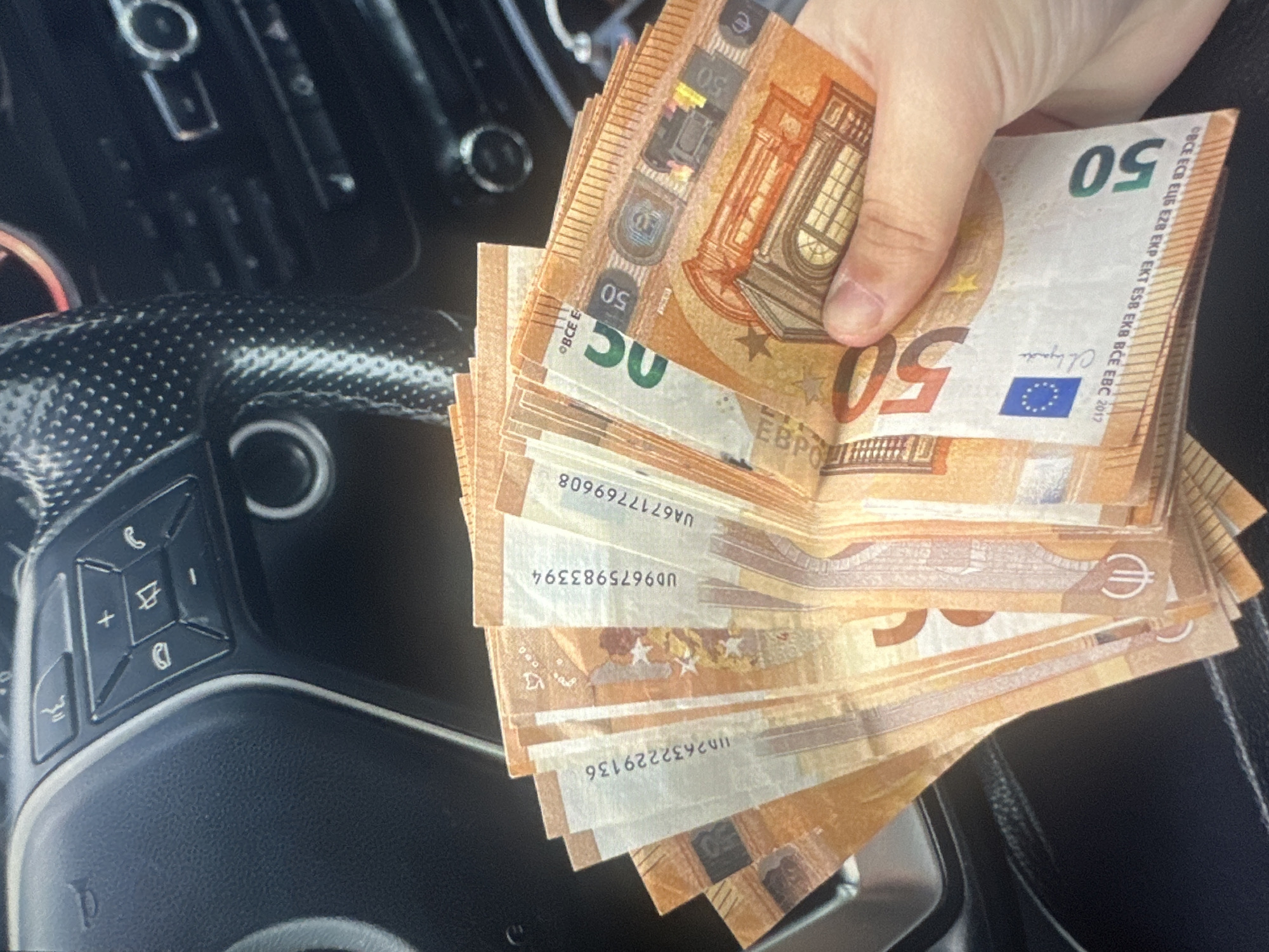 A close-up, high-angle shot shows a hand holding a fan of 50 Euro banknotes. The banknotes are predominantly orange and yellow, featuring architectural imagery and denomination numbers. The background is blurred, but appears to be the interior of a car, with a dark steering wheel and dashboard visible. The steering wheel has several buttons, including a phone icon and volume controls. The image does not provide any information or visual cues to suggest it is located in Tallinn, Estonia, or any other specific city.