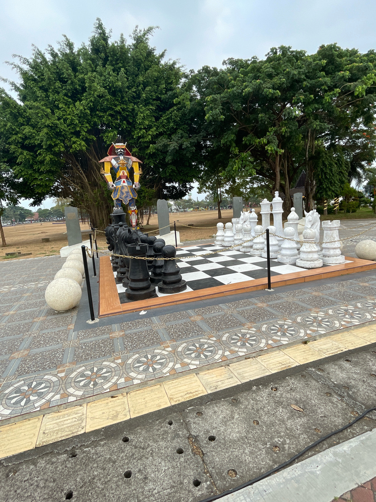 The image depicts a large, outdoor chess set located in Purworejo, Indonesia. The chess board features alternating black and white squares, with large, white chess pieces on one side and black pieces on the other. Behind the chess set, a colorful statue of a robot-like figure stands tall, partially obscured by the lush green foliage of a large tree. The area is paved with decorative tiles, and a chain fence surrounds the chess set. In the background, a wide, open field with scattered trees suggests a park or public space. The sky is overcast, indicating a cloudy day.